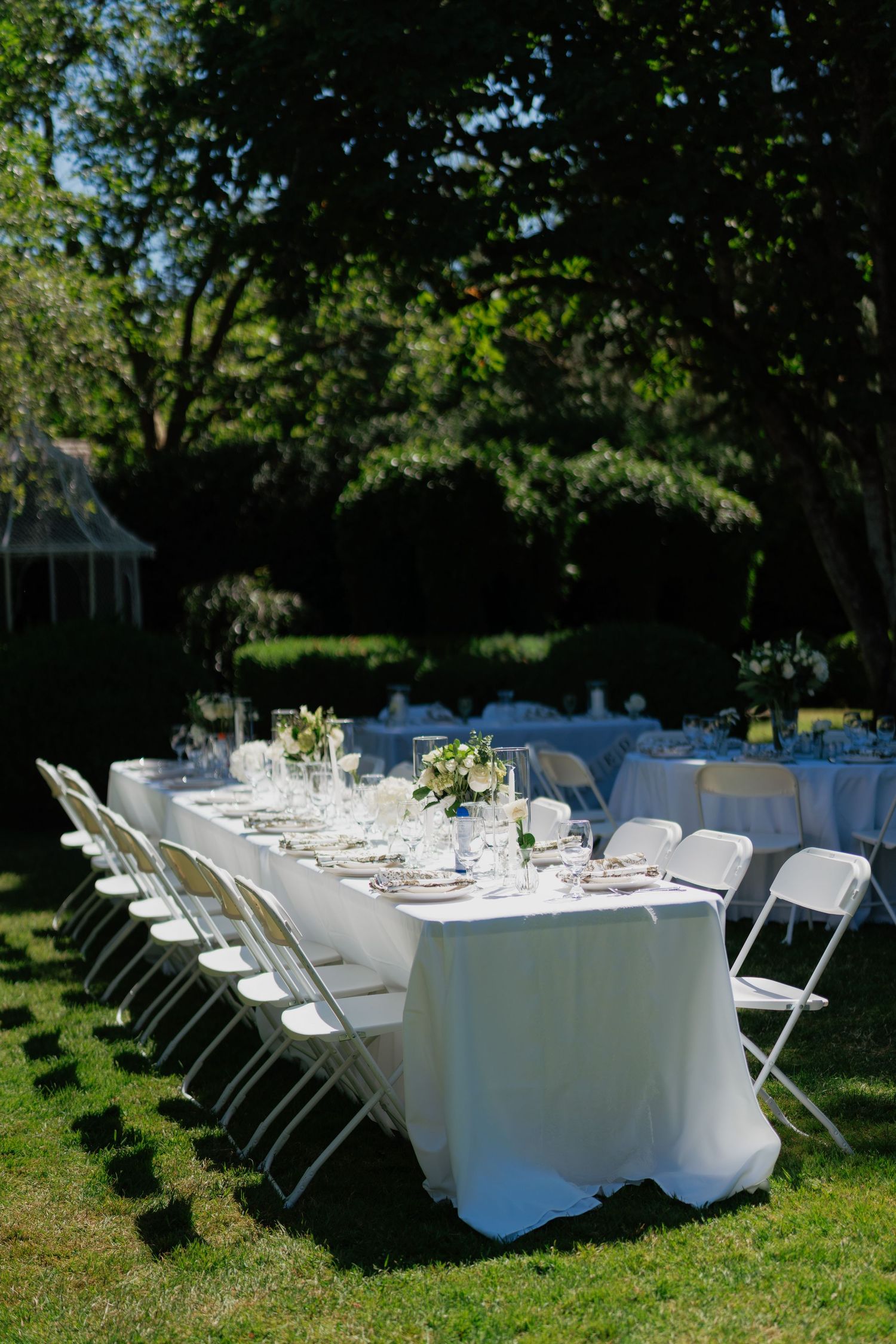 Elegant outdoor wedding reception with long white-draped tables and folding chairs set on lush green lawn surrounded by trees.