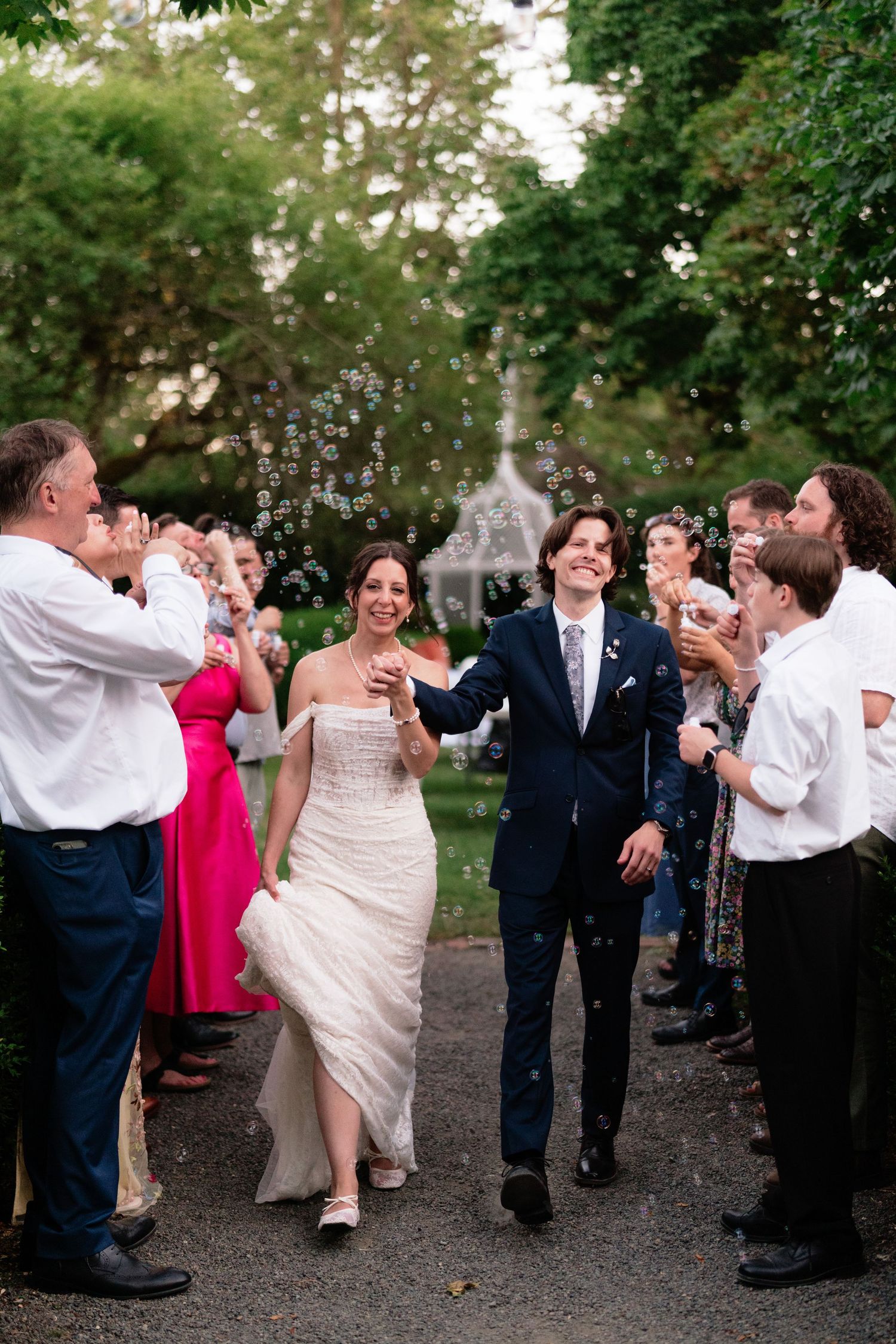 Joyful newlyweds walk through bubble send-off surrounded by cheering guests on garden pathway after outdoor wedding.