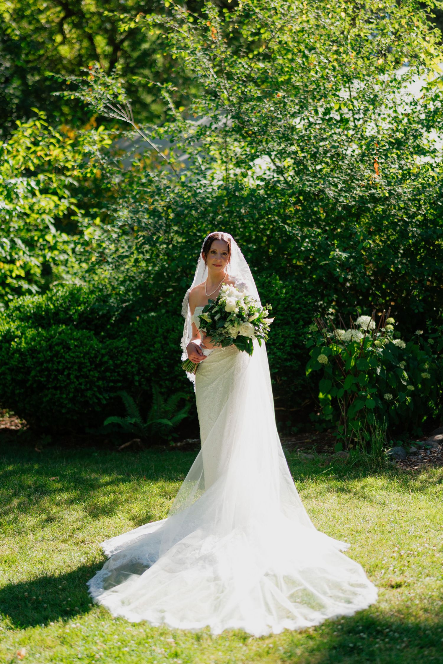 Elegant bride in a white lace wedding gown with cathedral veil holding a bouquet in a sunlit garden.