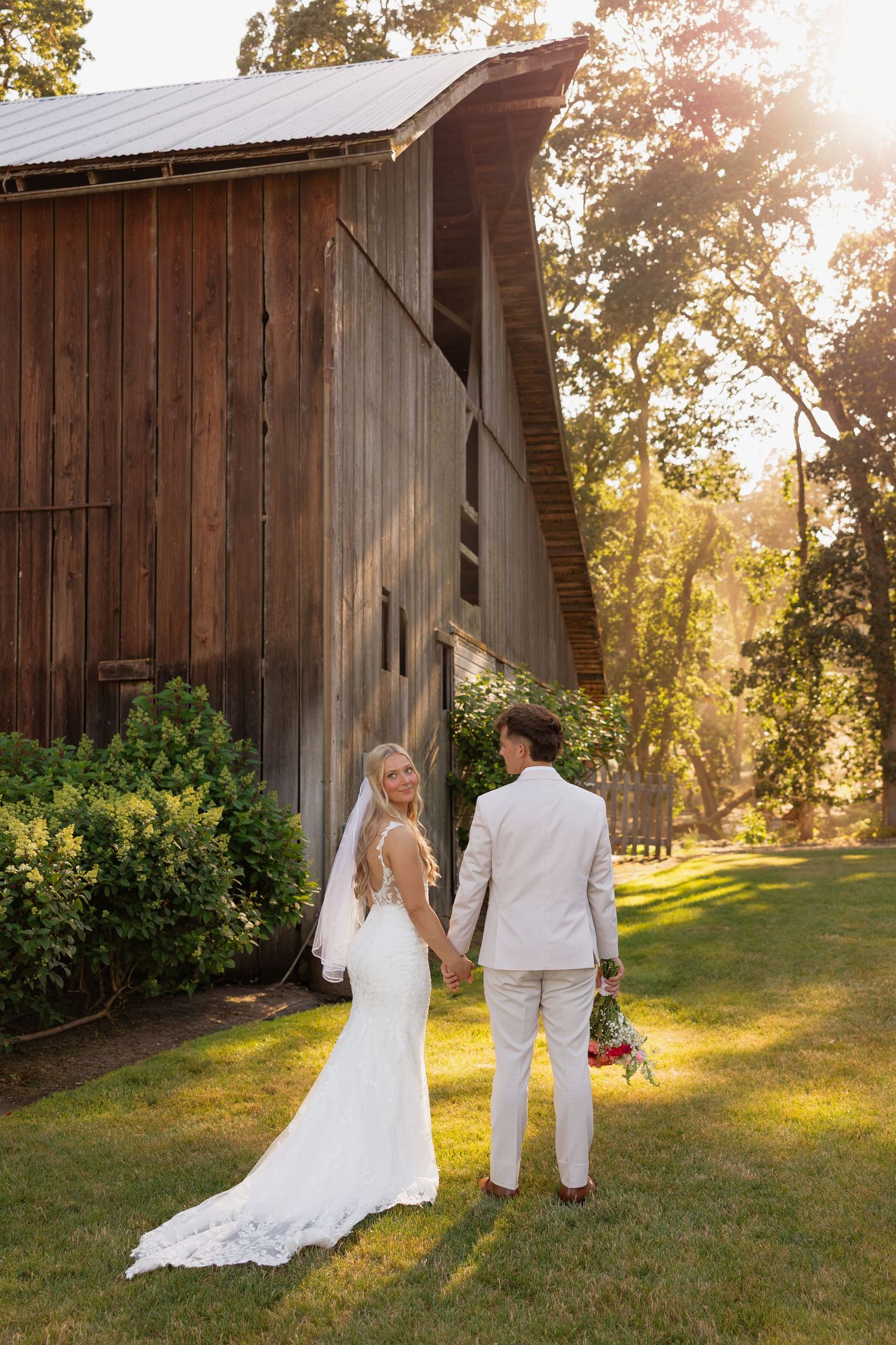 Bride and groom hold hands at golden hour beside a rustic wooden barn, bride in lace gown with veil.