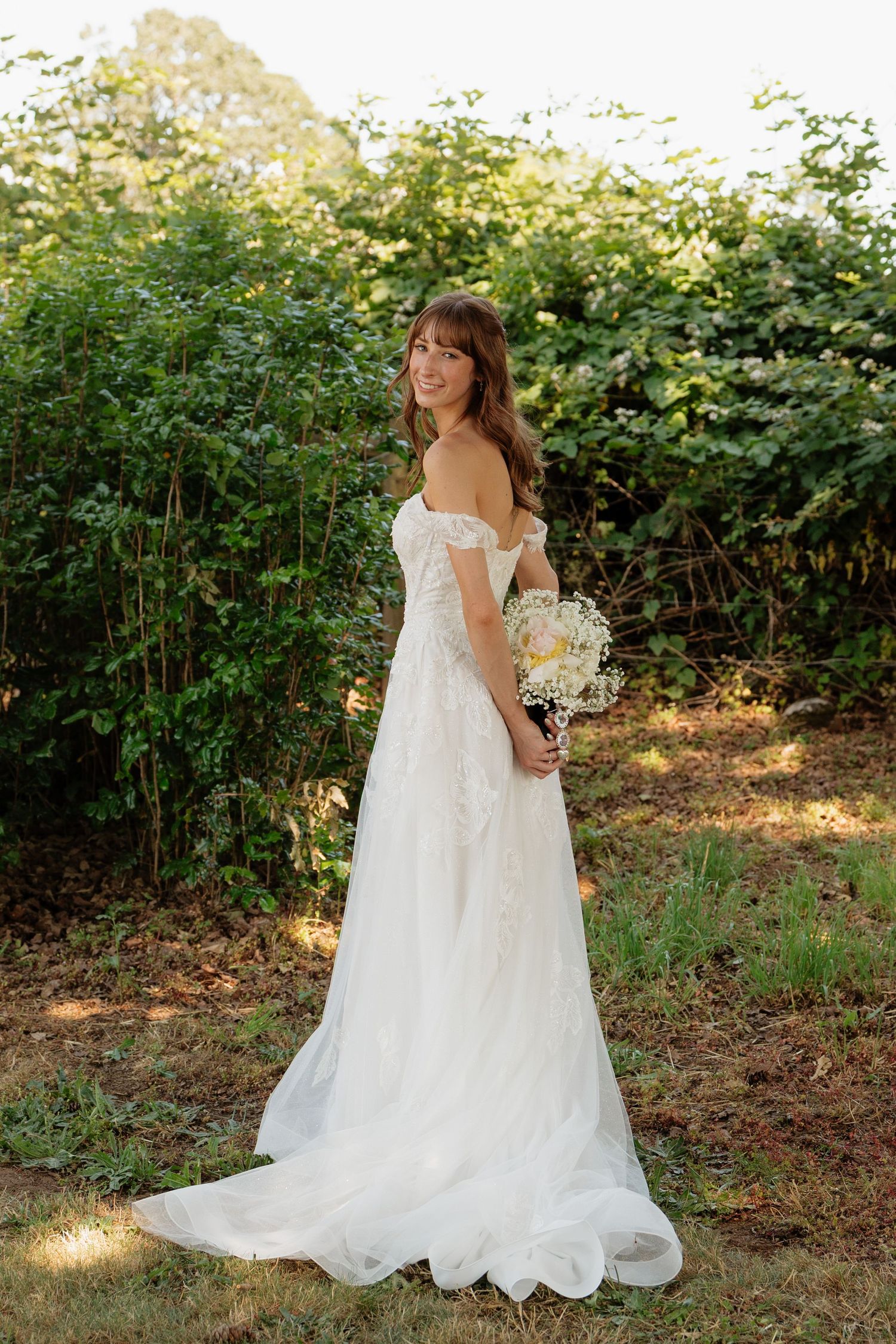 Smiling bride in off-shoulder white lace gown holding bouquet, posing outdoors among lush green bushes.