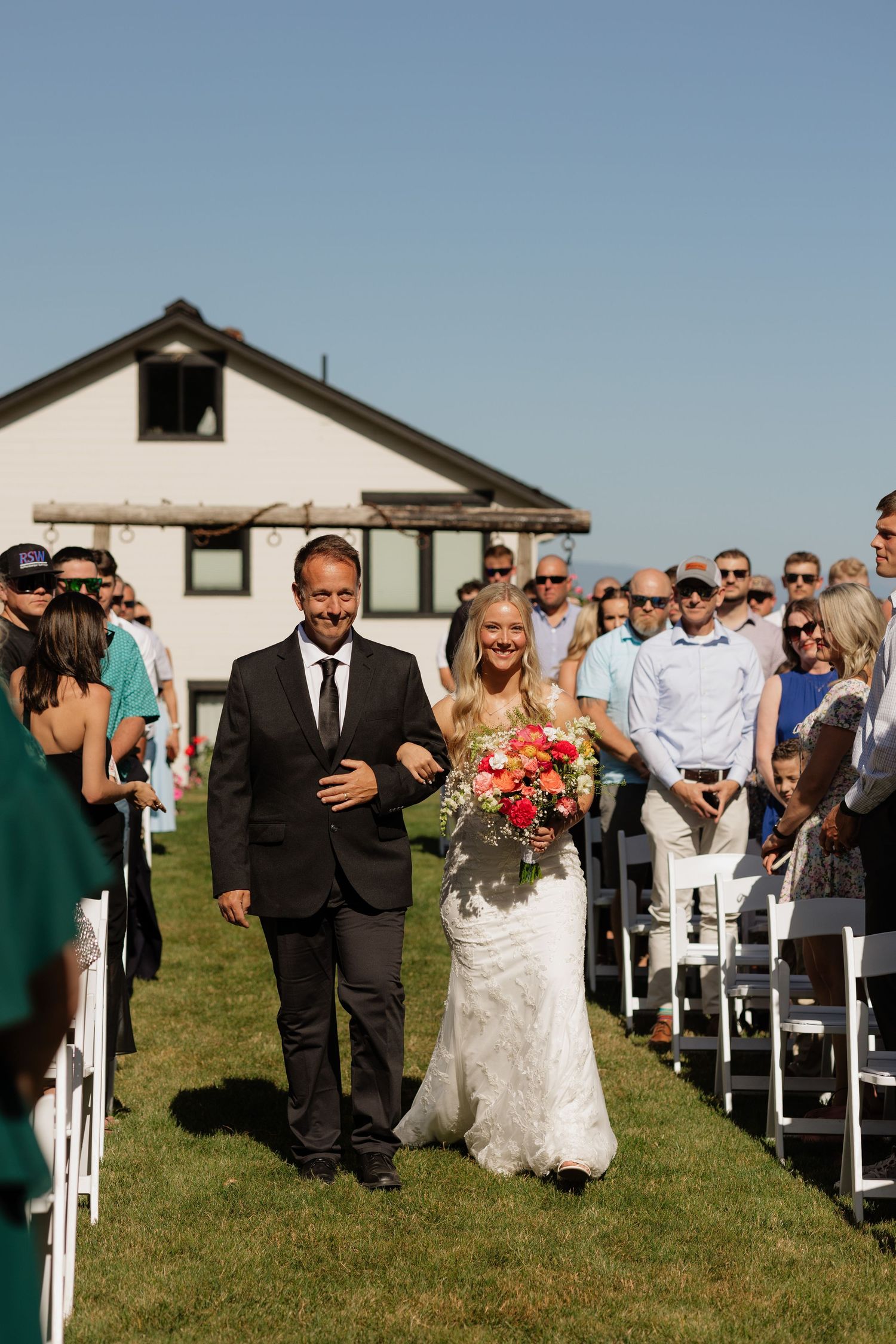Bride walks down outdoor wedding aisle with escort, holding vibrant bouquet, guests seated on white chairs.