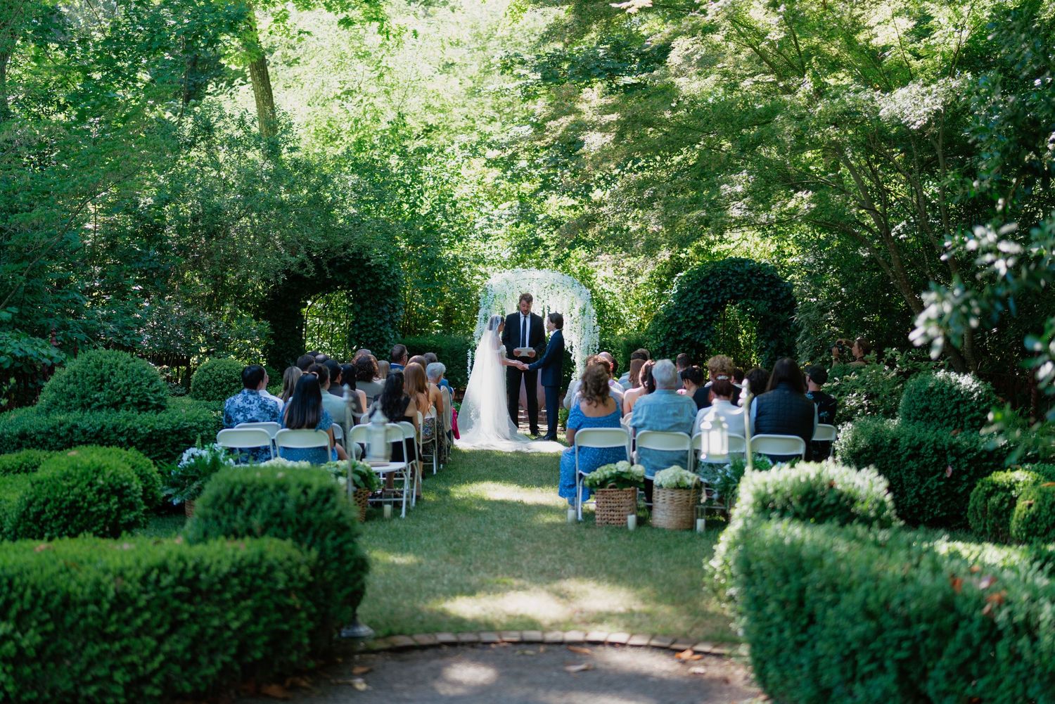 Outdoor garden wedding ceremony with guests seated along lush green hedgerows.