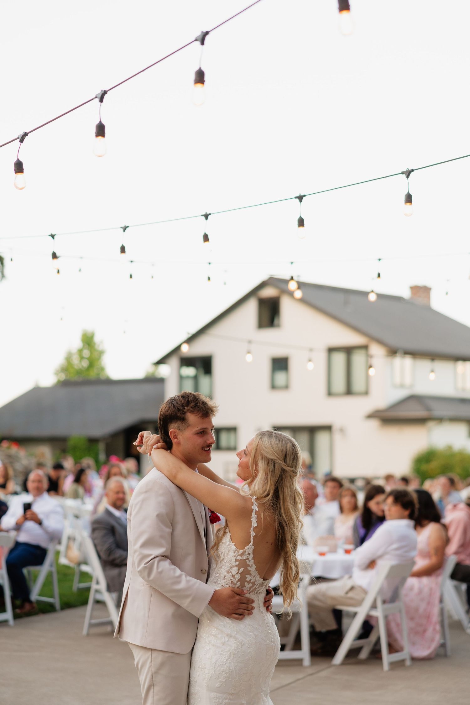 Bride and groom sharing first dance outdoors under string lights with wedding guests seated nearby.