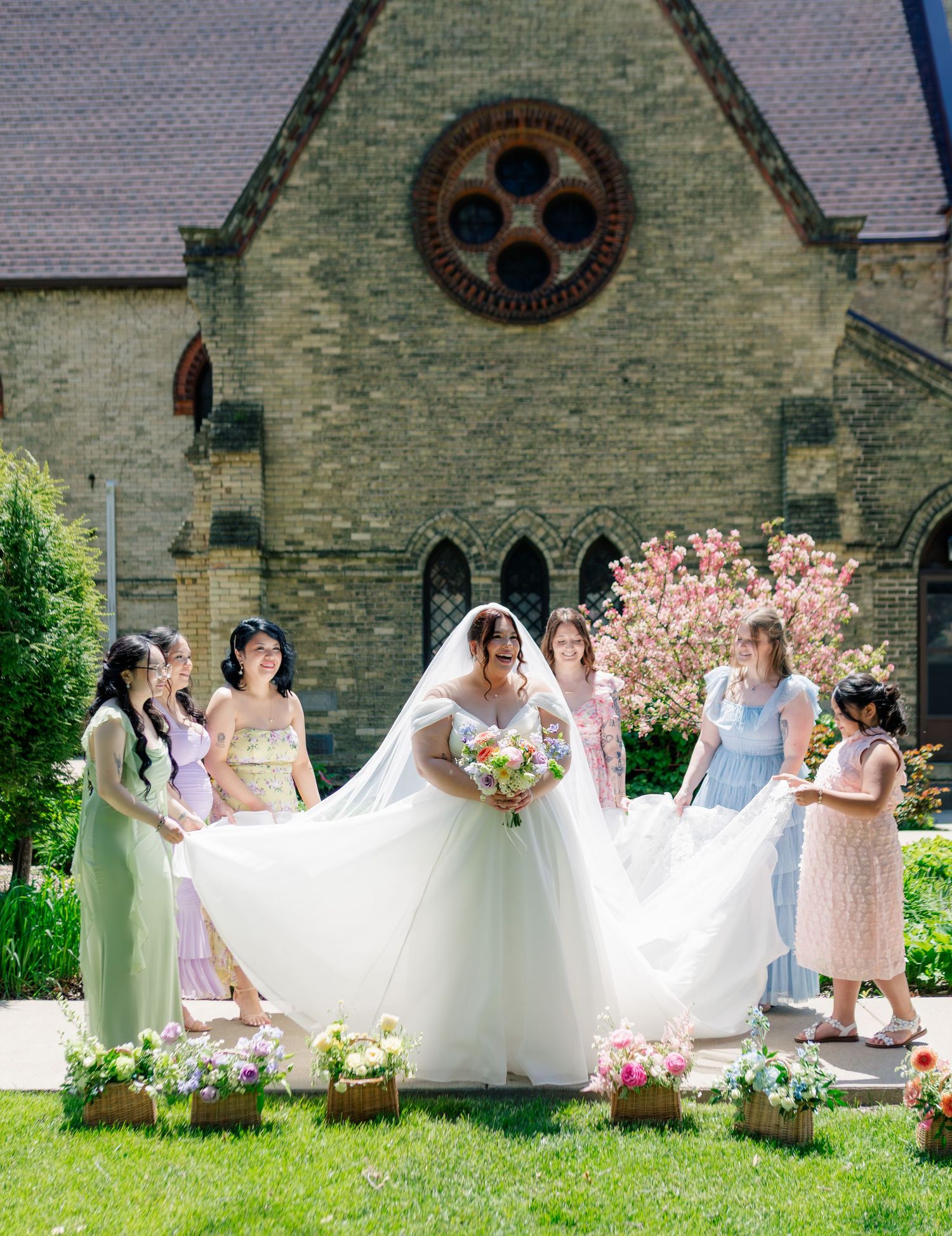 A wedding party gathers on a church lawn with bridesmaids in pastel dresses helping with the bride's flowing white gown.