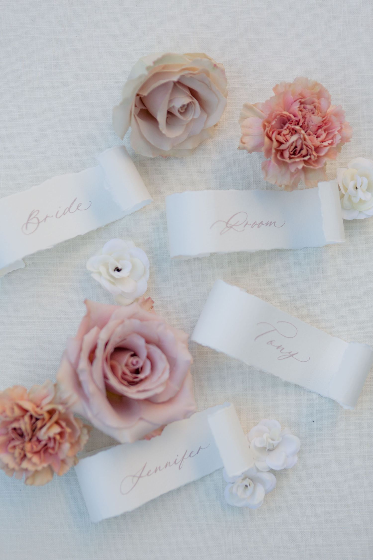 Elegant wedding place cards with calligraphy text surrounded by soft pink and white roses and carnations.