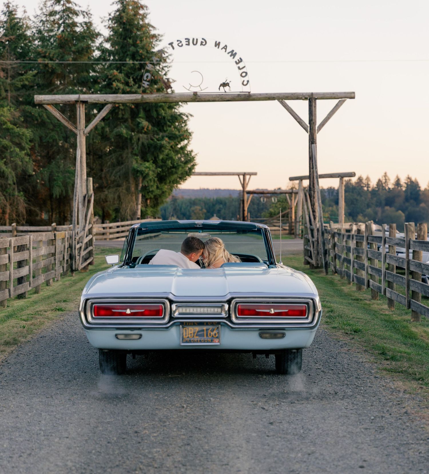 A classic Ford Thunderbird convertible parked on a gravel road under a wooden ranch entrance gate at sunset.