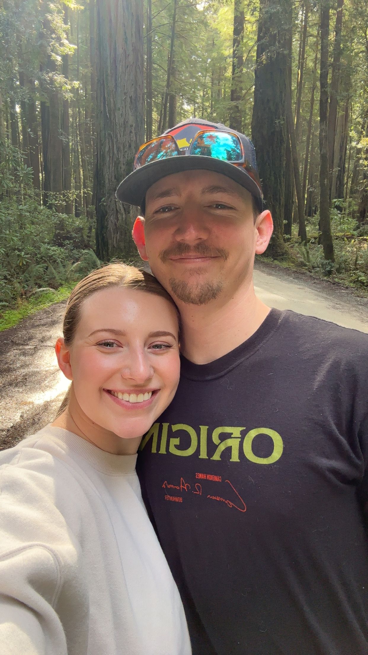 Couple smiling together on a forest trail surrounded by tall redwood trees.