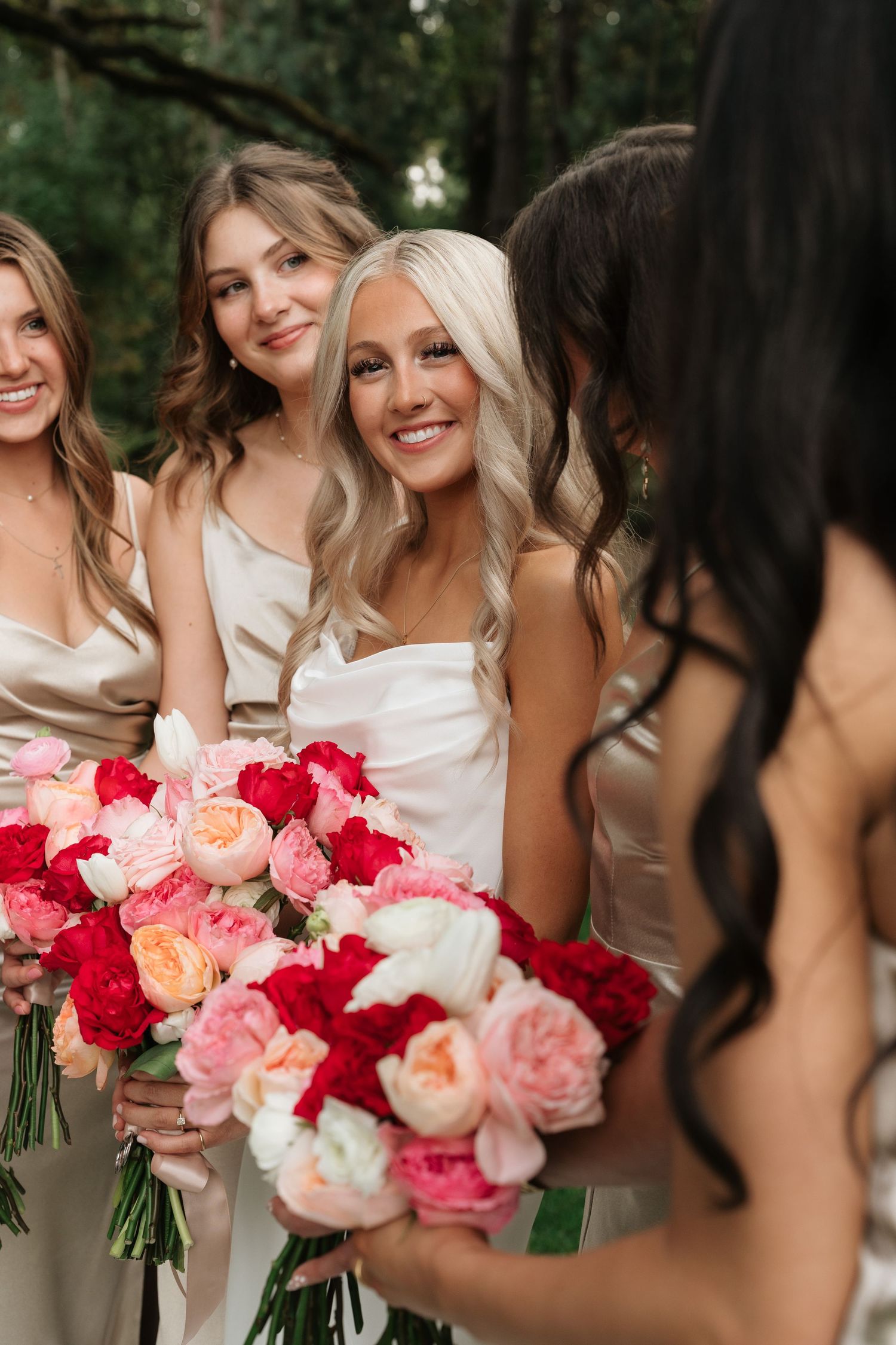 Bride in white strapless gown smiles with bridesmaids in champagne dresses holding red and pink peony bouquets.