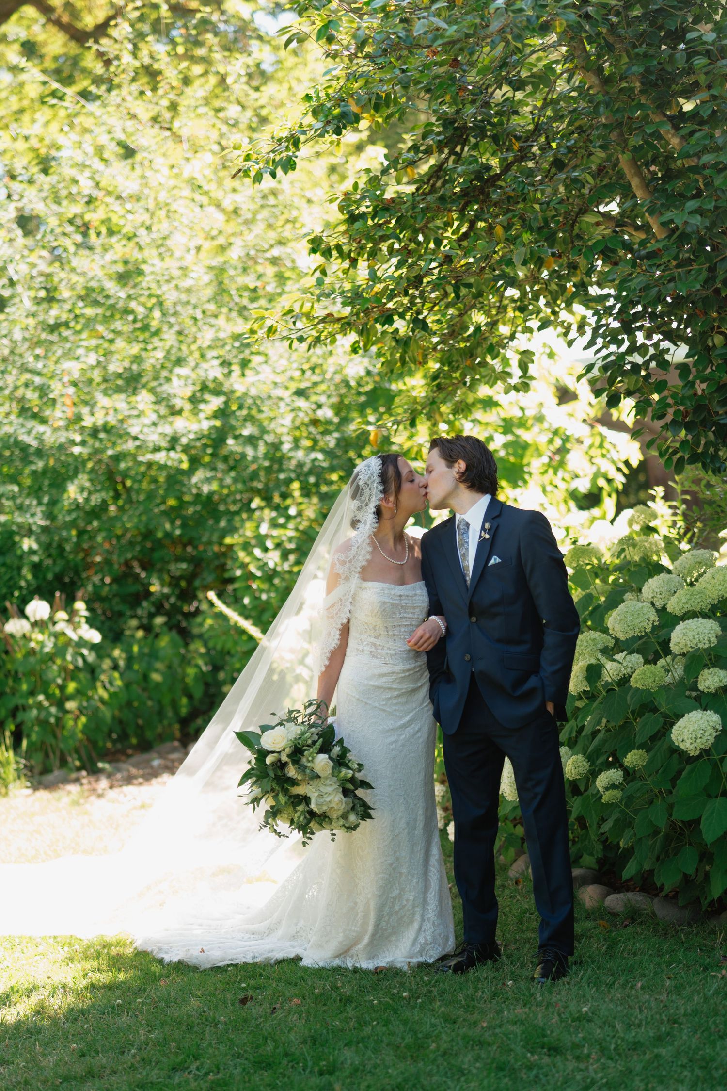 Bride and groom sharing a romantic kiss in a lush green garden, bride wearing a lace gown with veil.