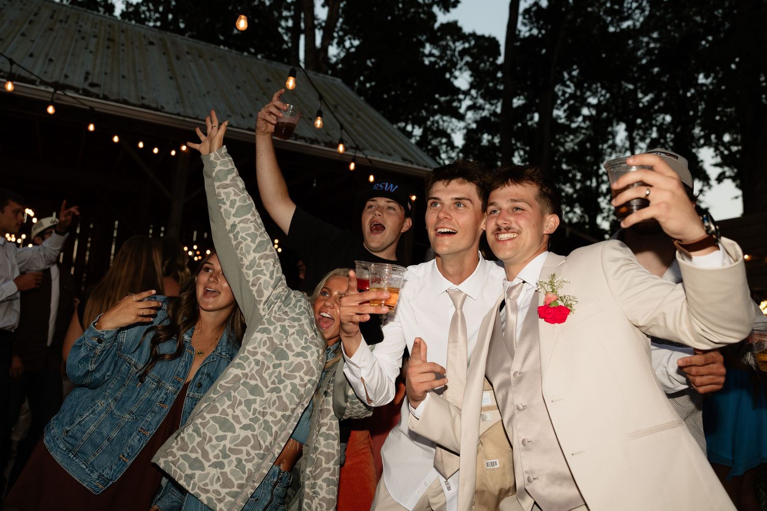 Groom in beige suit celebrating with wedding guests taking selfie under string lights at reception.