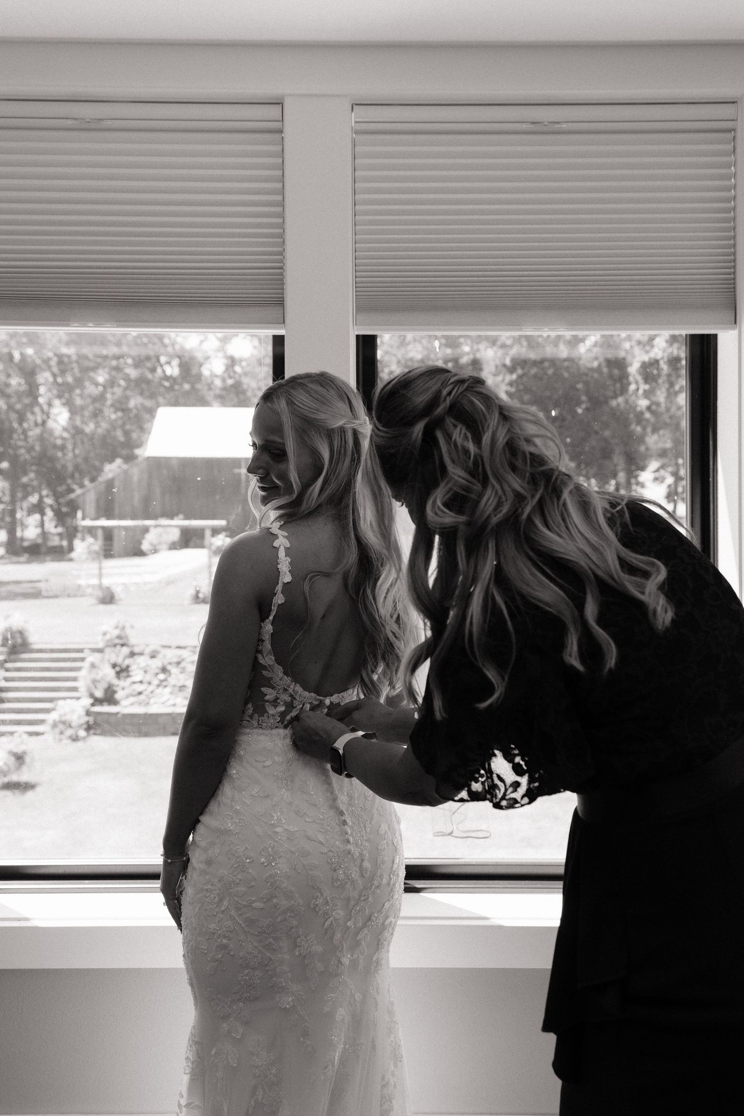 Black and white photo of a bridesmaid buttoning the back of a brides lace wedding dress by a window.