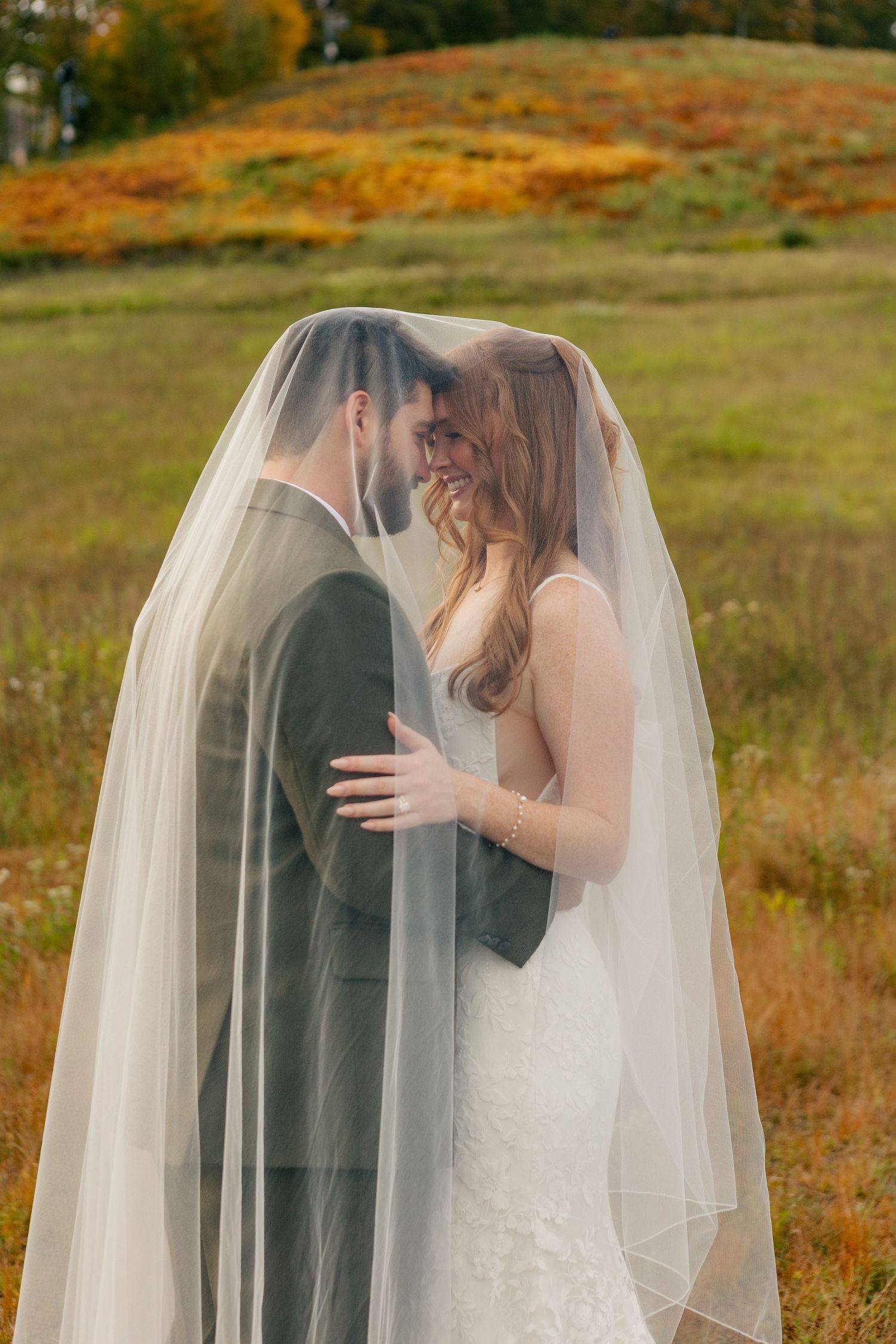Bride and groom share an intimate moment under a sheer veil in an autumn field with colorful foliage.