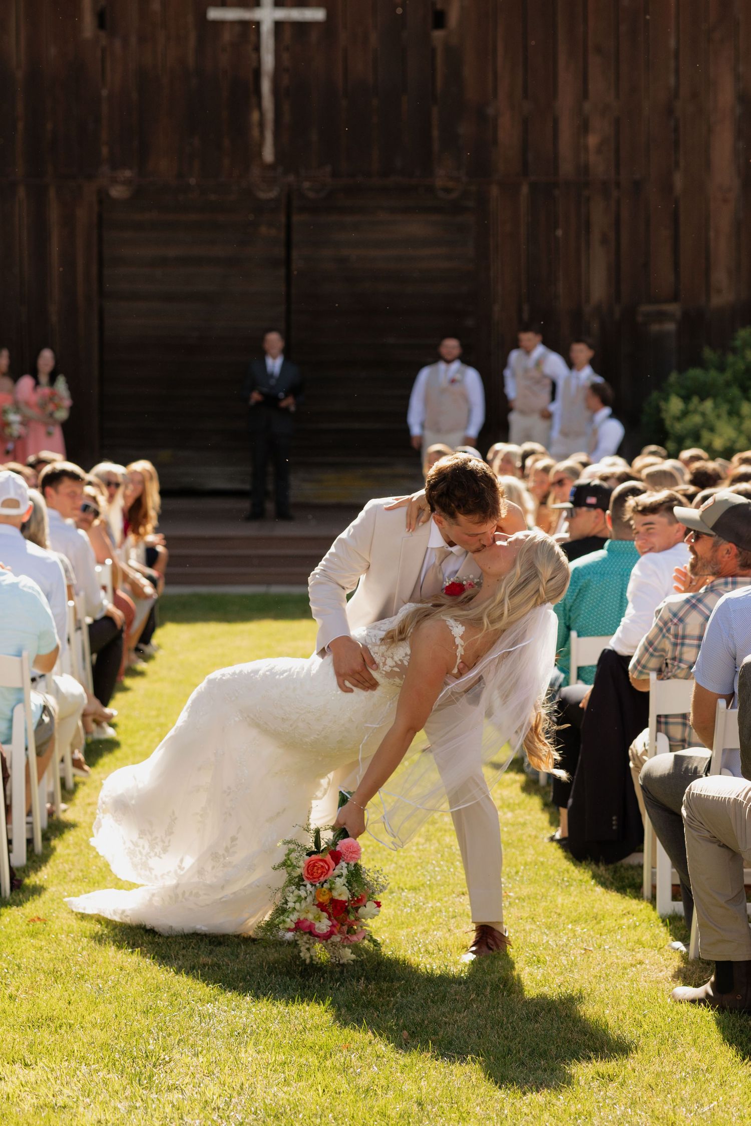 Bride and groom sharing romantic dip kiss at outdoor wedding ceremony aisle with guests watching.