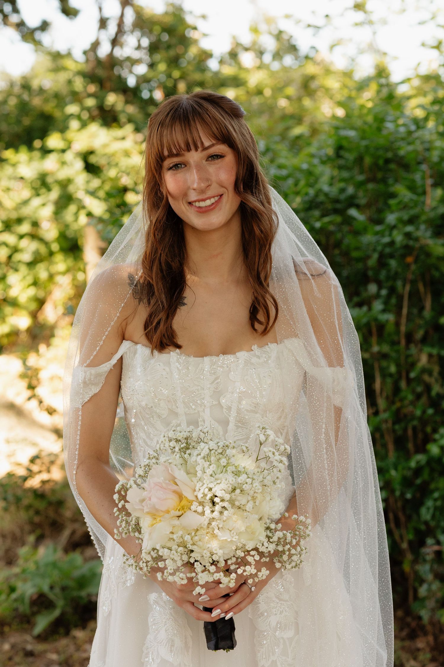 Smiling bride in lace gown and veil holds a white floral bouquet outdoors in natural sunlight.