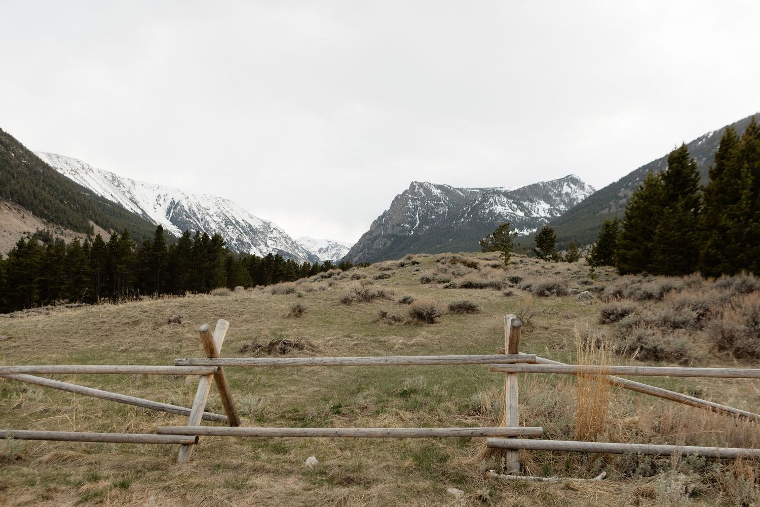 Rustic wooden fence overlooking a mountain valley with snow-capped peaks and evergreen trees under an overcast sky.
