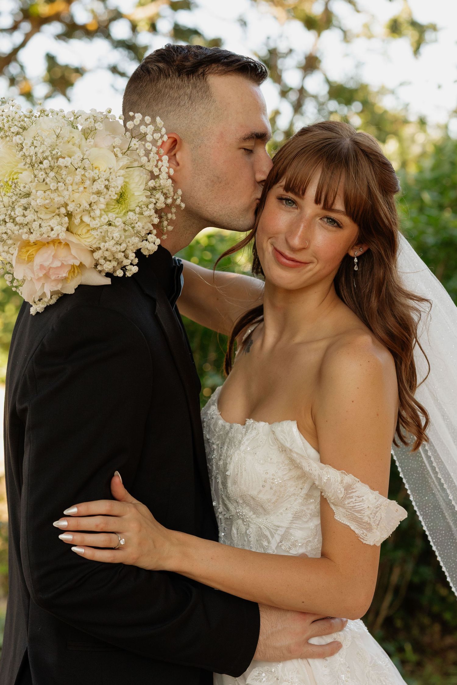 Groom kisses bride on cheek as she smiles softly, holding bouquet with peony and baby's breath.