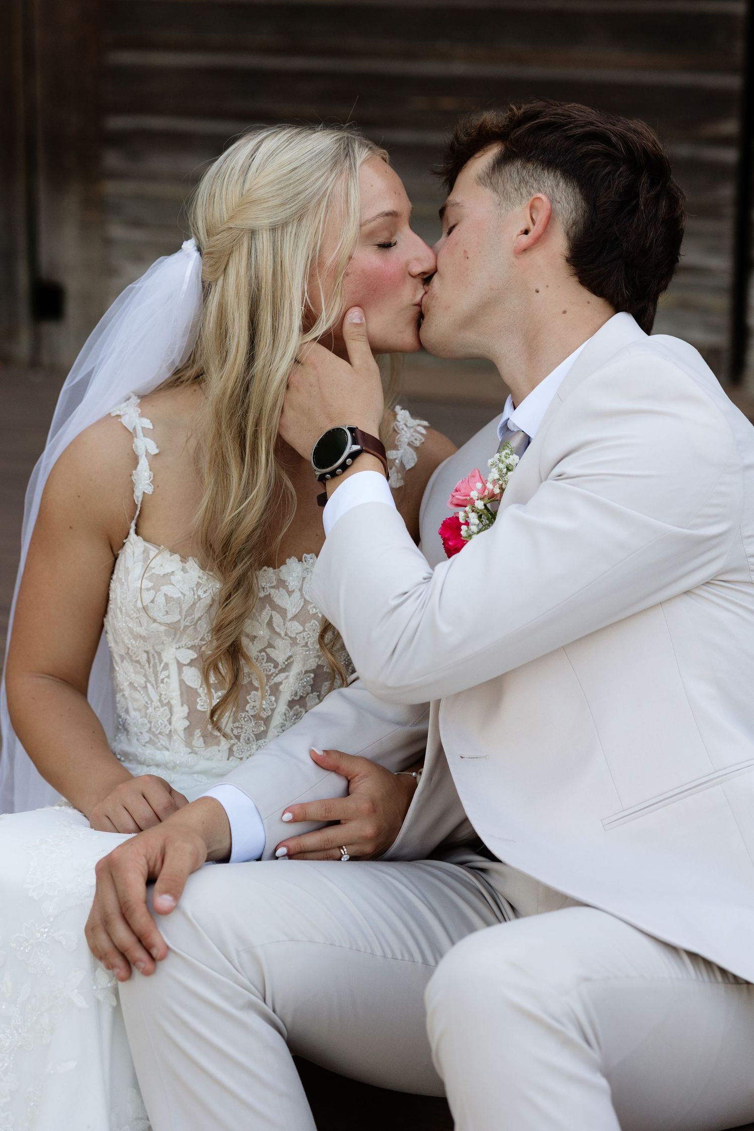 Bride and groom share tender kiss while seated together on their wedding day outdoors.