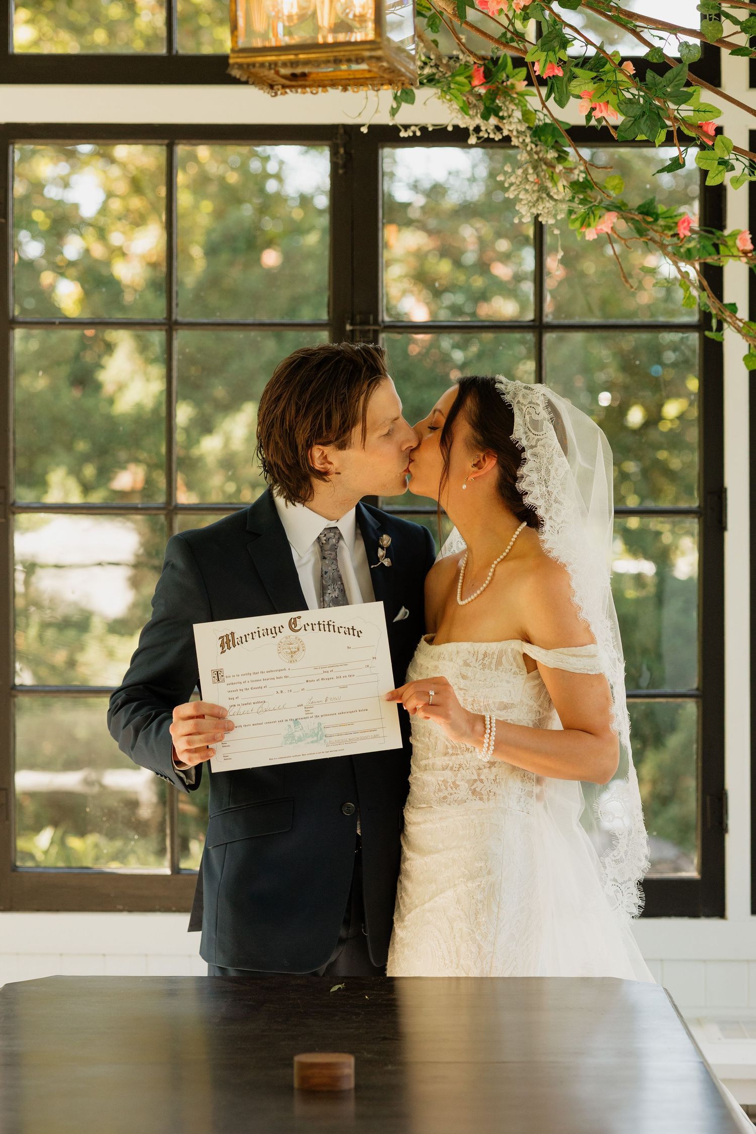 Newlyweds kissing while holding their marriage certificate in front of large windows at their wedding venue.