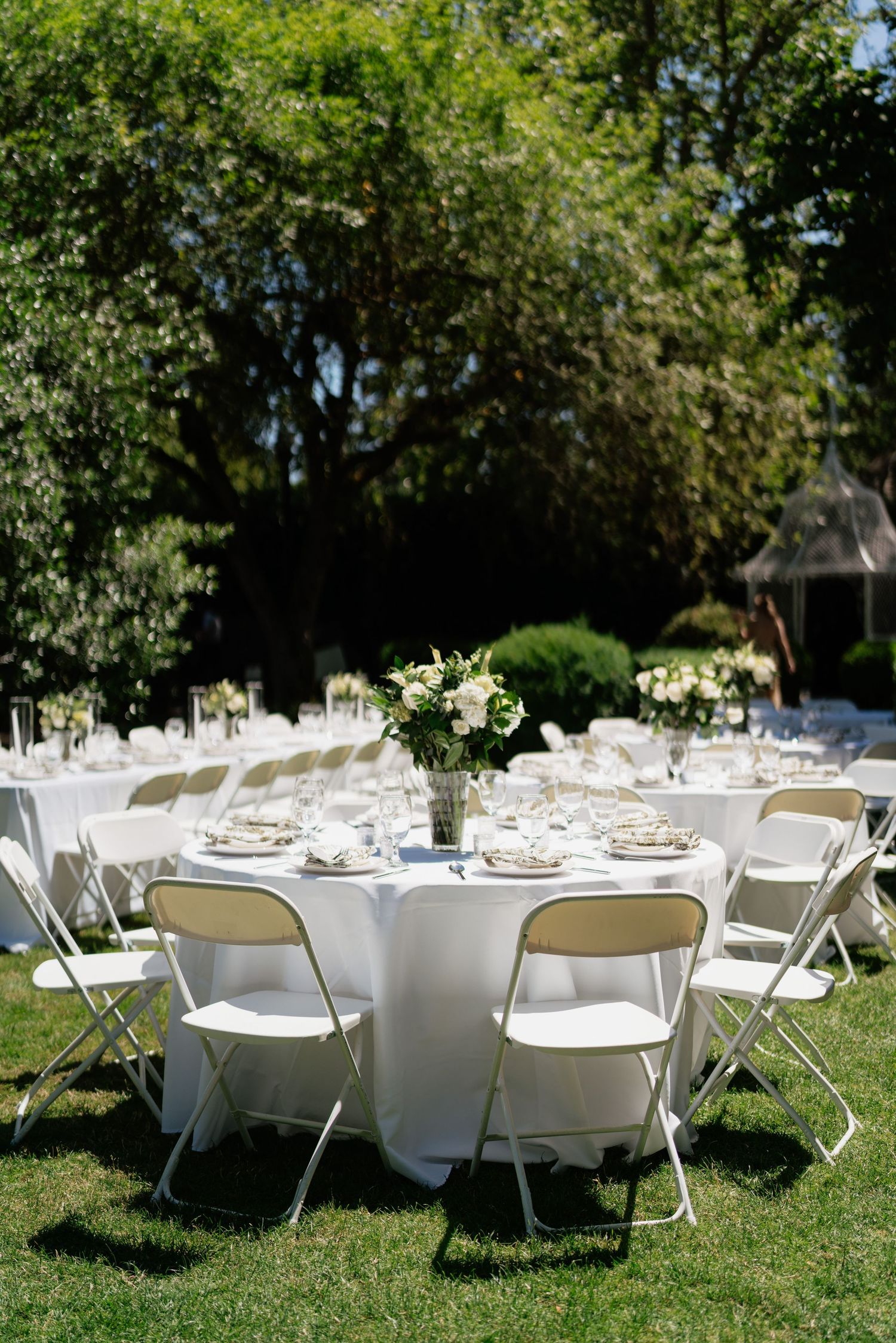 Elegant outdoor wedding reception with round tables draped in white linens, floral centerpieces, and white folding chairs on green lawn.