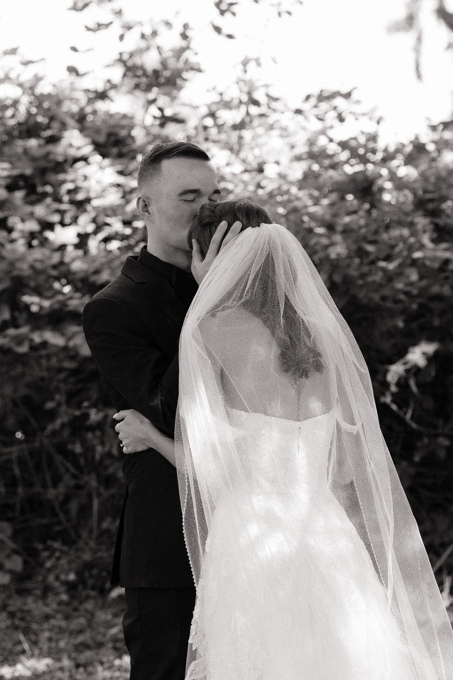 Black and white photo of groom kissing bride in white veil and gown outdoors after wedding ceremony.
