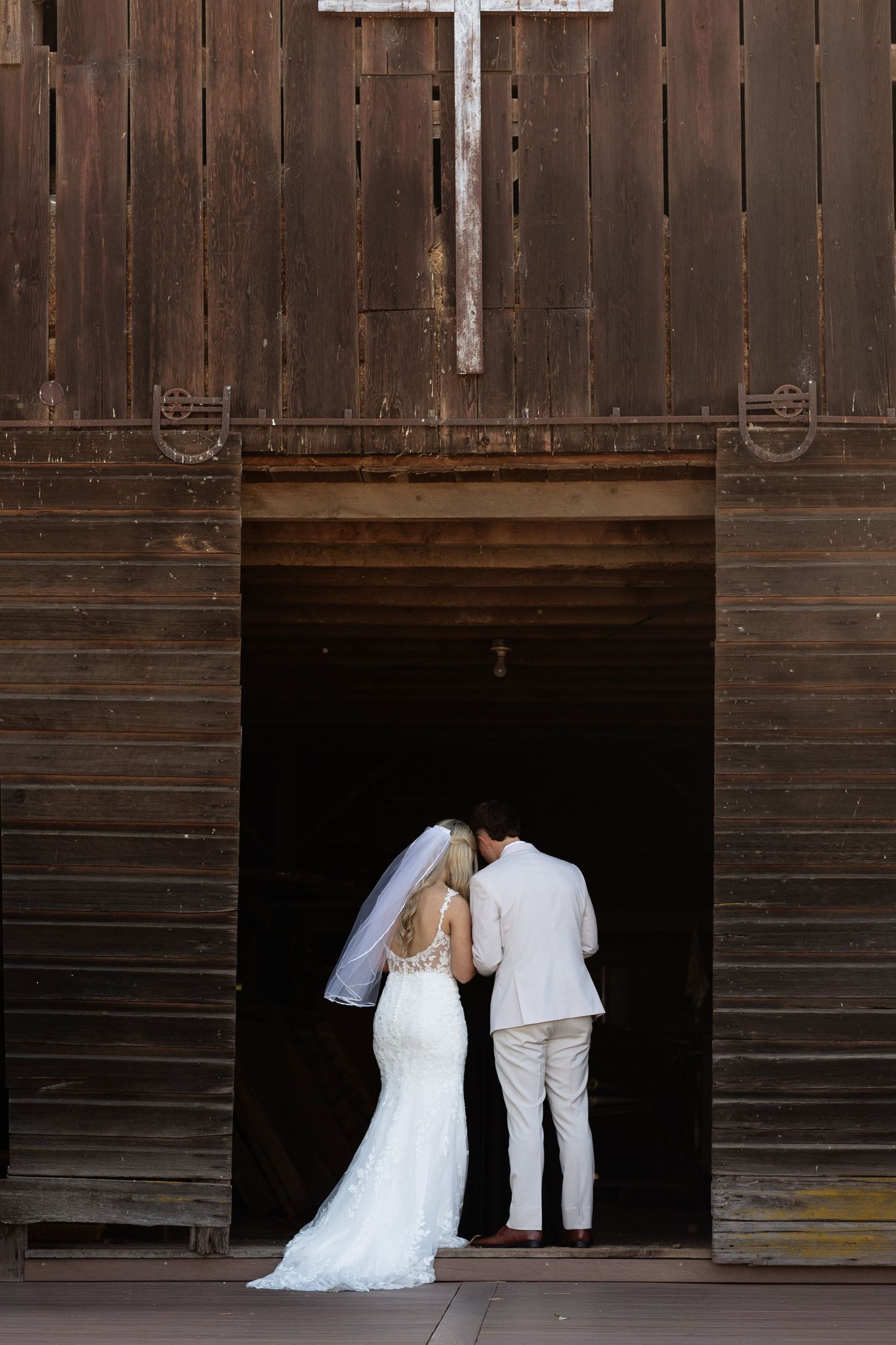 Bride and groom sharing intimate moment in rustic wooden barn doorway with white cross above.