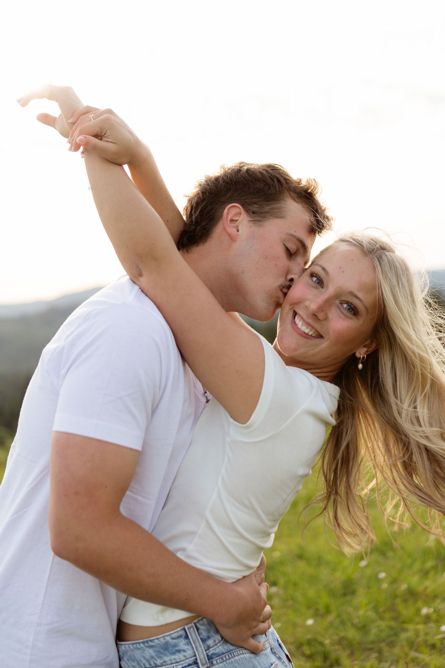 Young couple in white shirts sharing a romantic kiss outdoors in a sunny green meadow.