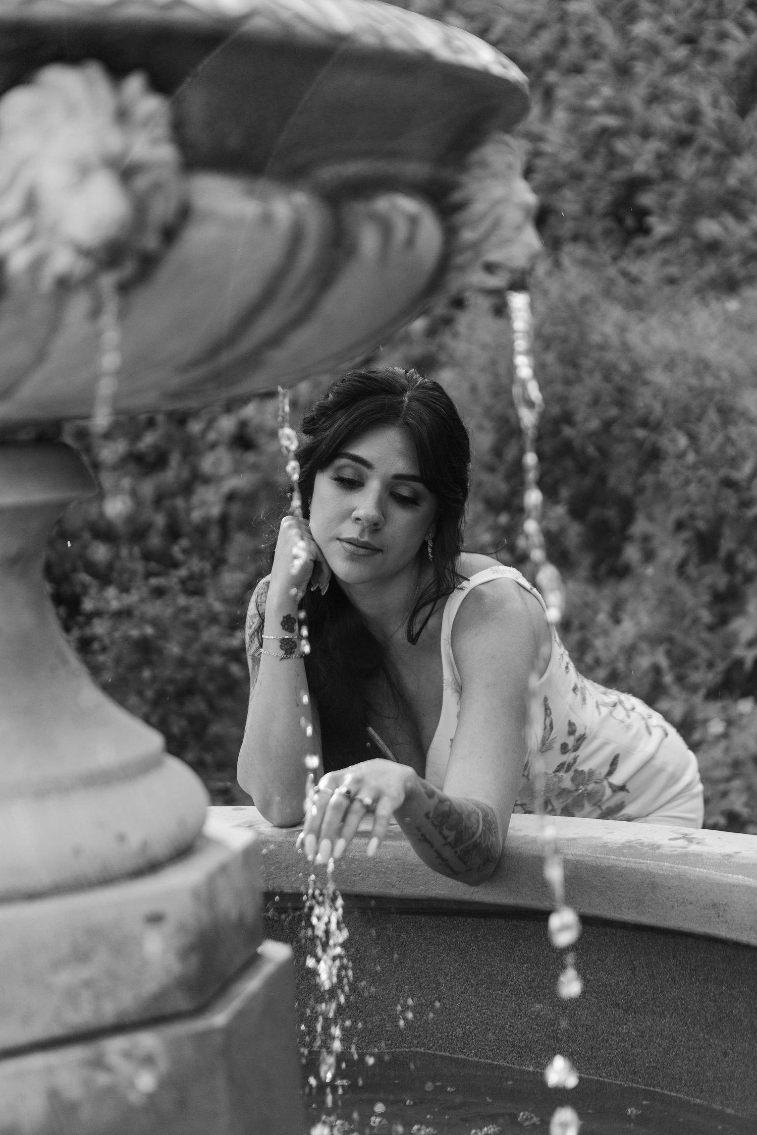 Black and white photo of woman in sleeveless top leaning on fountain with running water streams in garden setting.