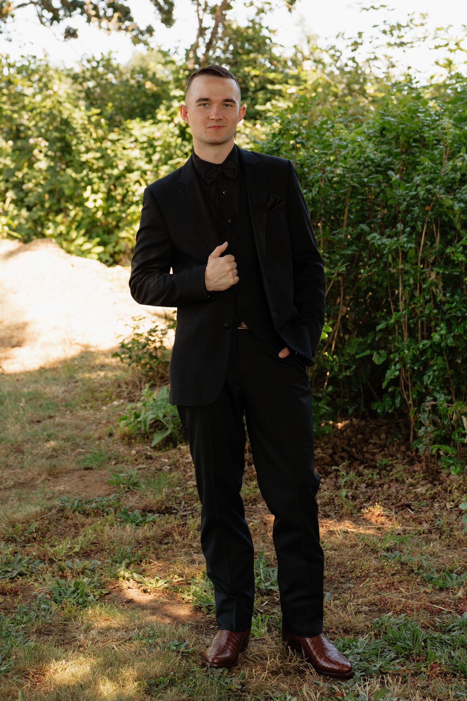 Groom in black suit and brown shoes posing confidently in a sunlit garden setting.