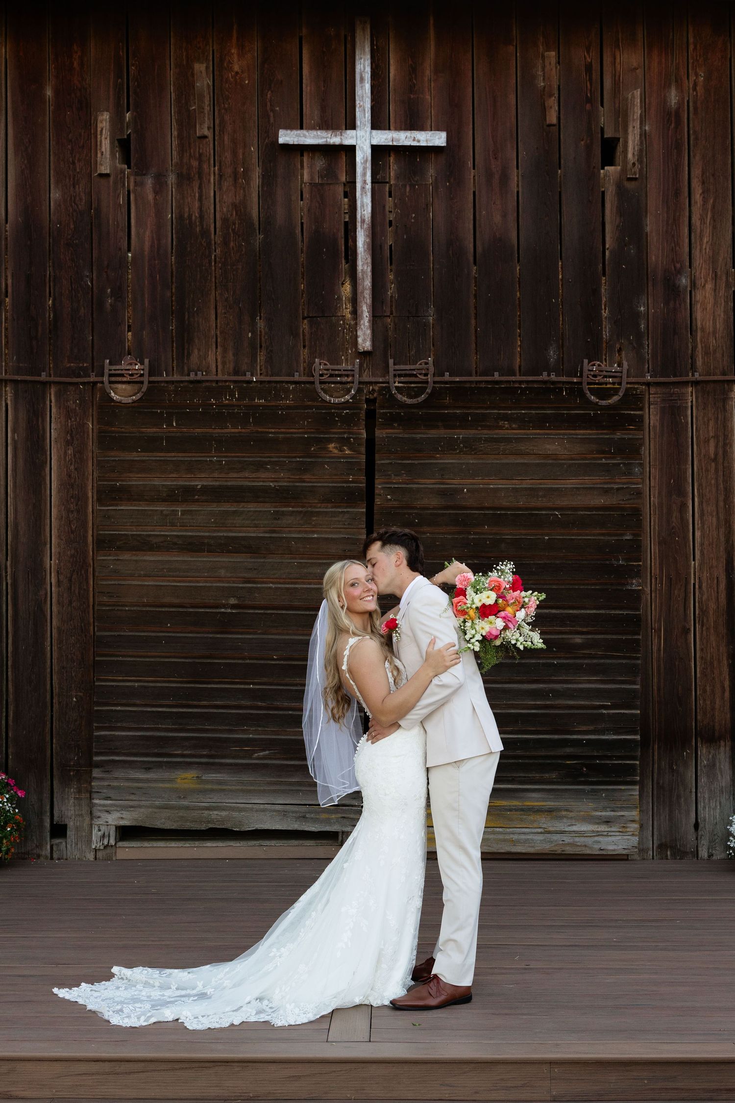 Bride and groom embracing in front of rustic barn doors with a white cross, bride holding colorful bouquet.