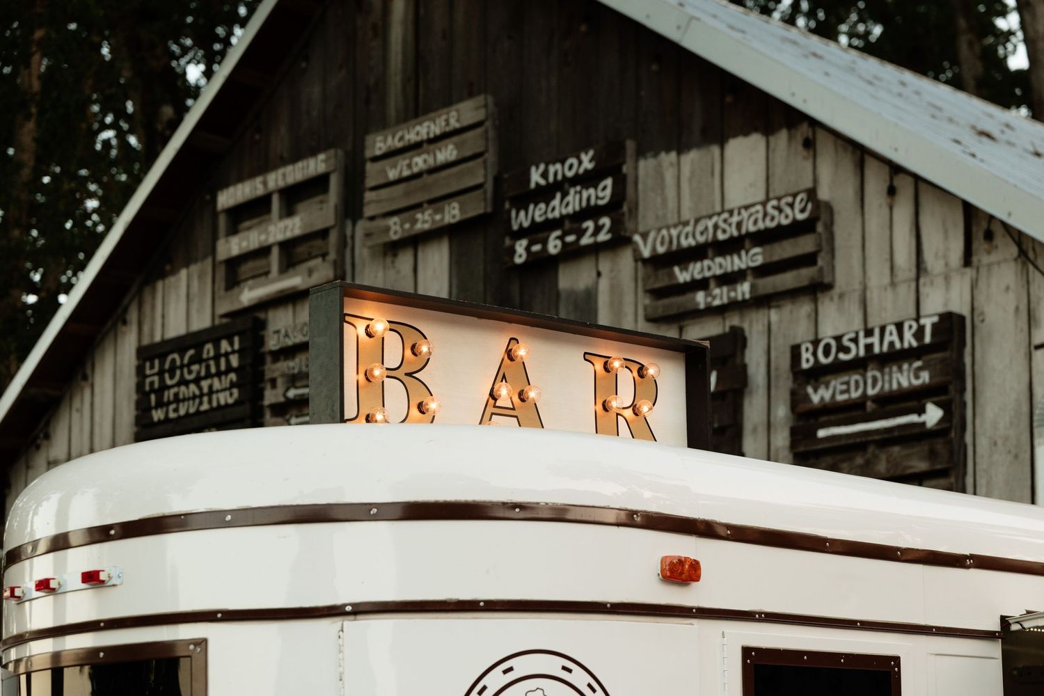 Vintage horse trailer converted into a wedding bar with illuminated BAR sign and rustic wooden directional signs.