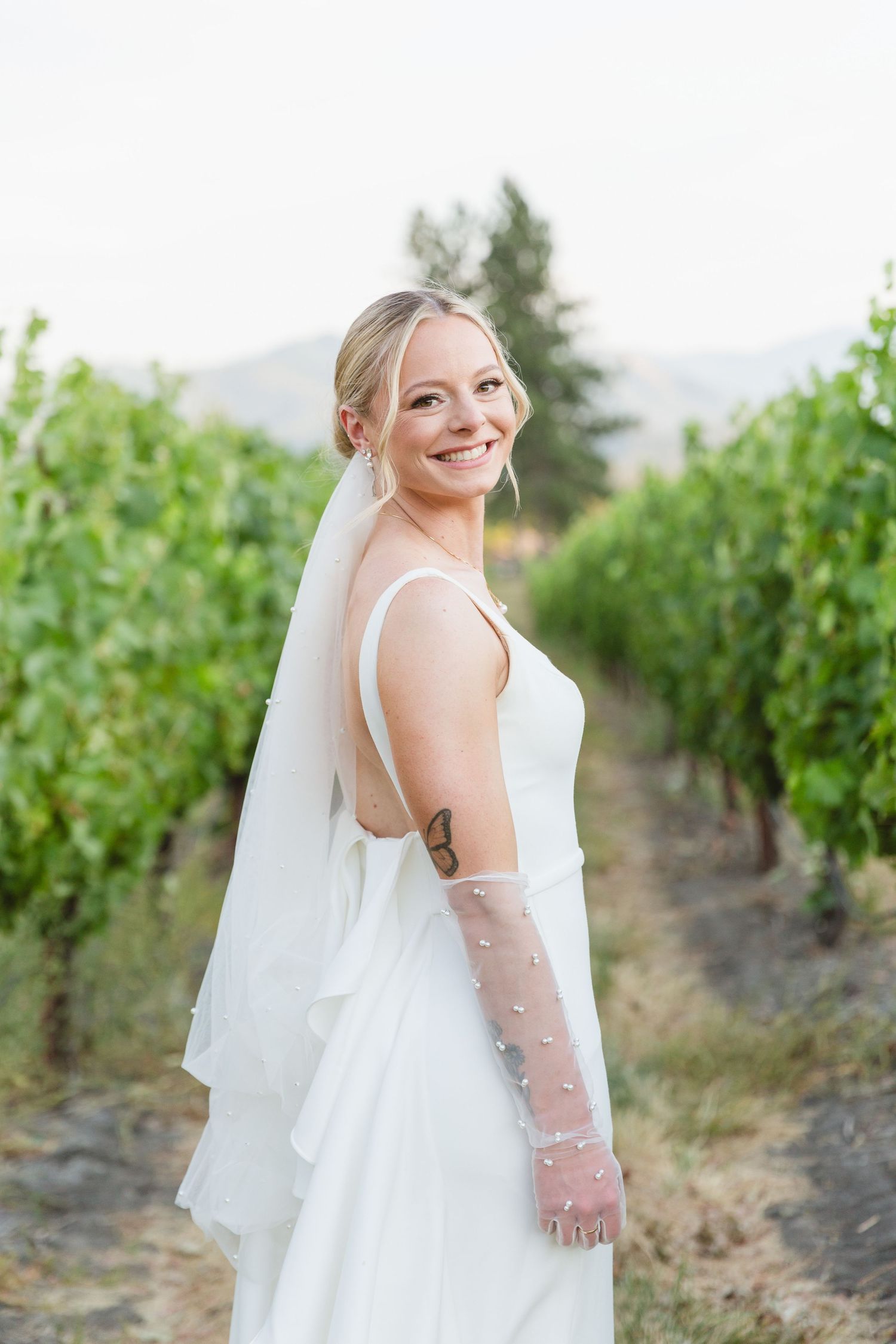 A bride in a sleeveless white wedding dress and veil poses in a scenic vineyard with mountains in the background.