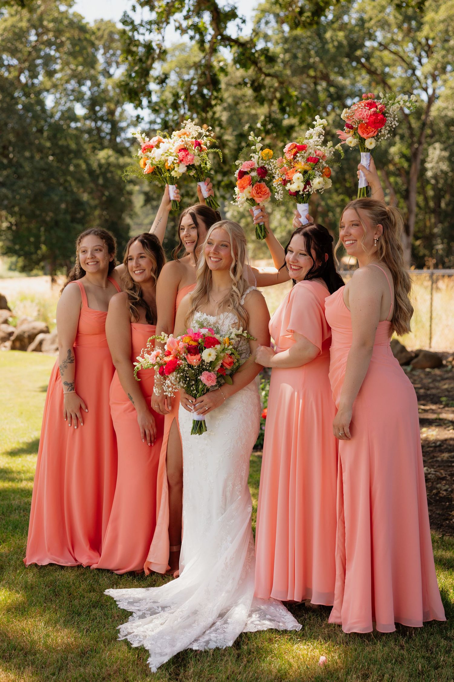Smiling bride and bridesmaids in coral gowns raise colorful bouquets joyfully outdoors on wedding day.