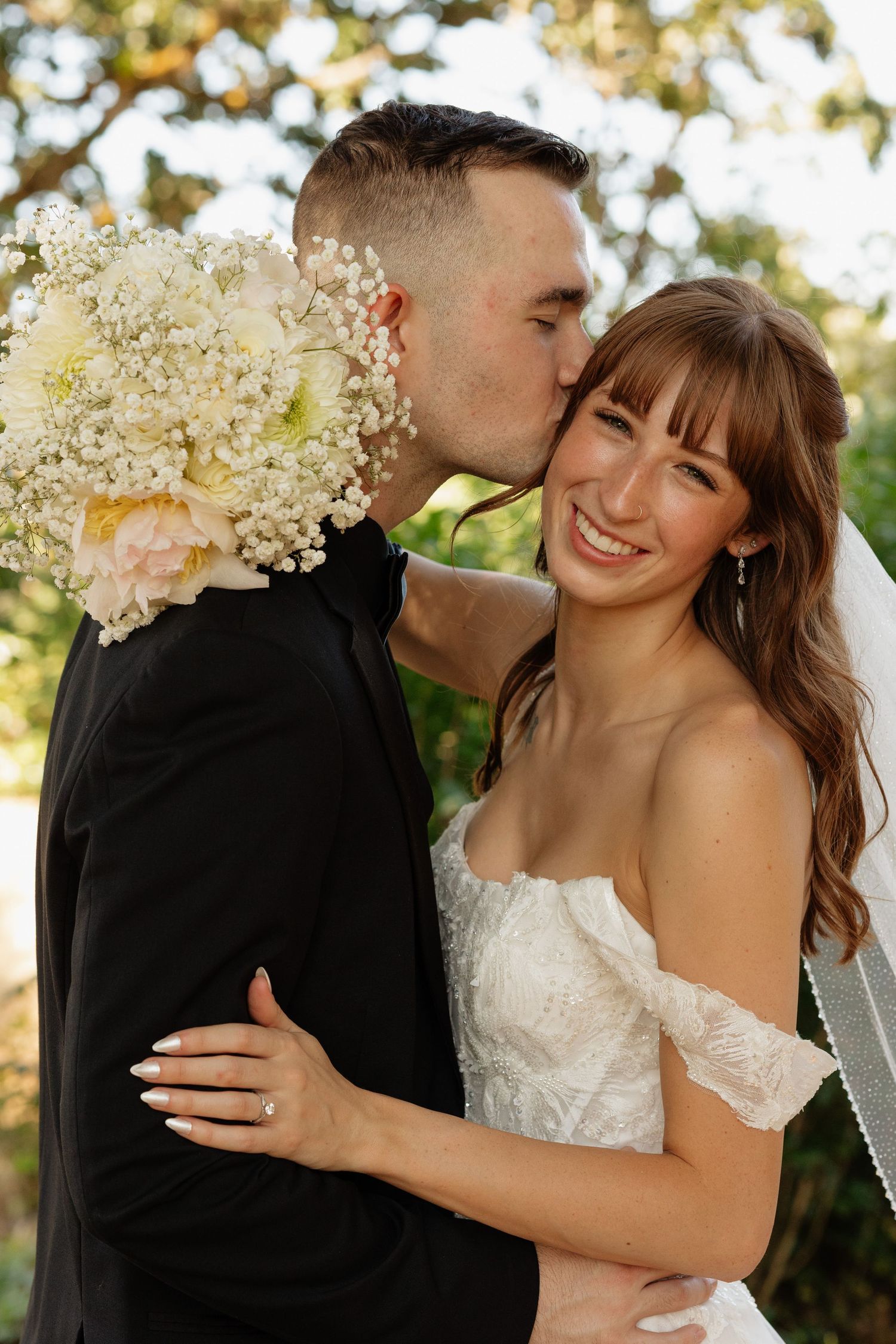 Groom kisses smiling bride on cheek outdoors, she holds white floral bouquet in natural light.