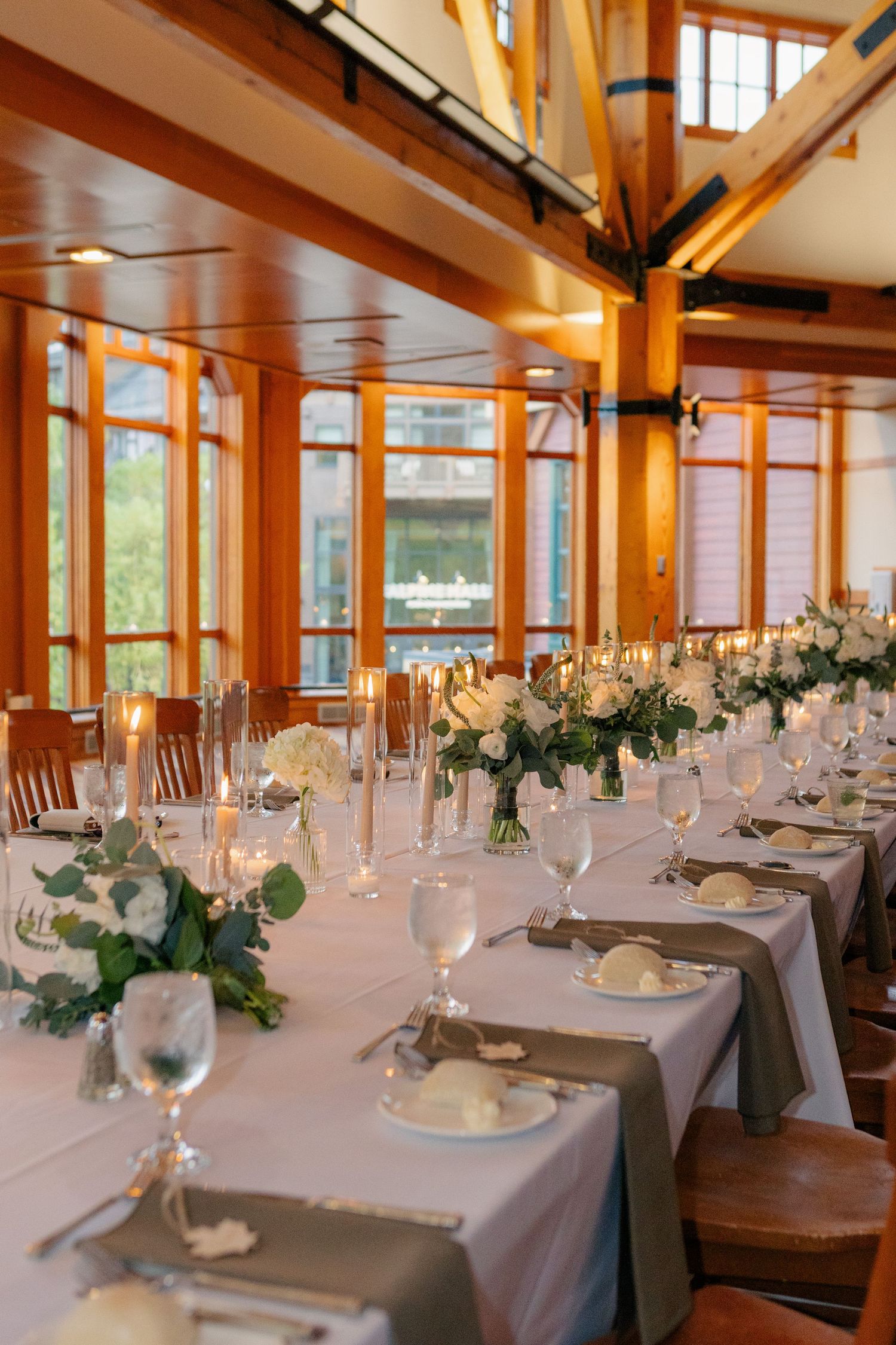 Elegant wedding reception hall with long white tables, floral centerpieces, candles, and warm wood-framed windows.