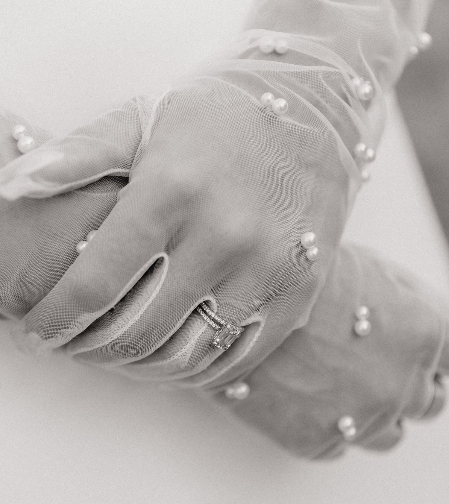 Black and white photo of hands wearing pearl-embellished tulle gloves with a sparkling diamond engagement ring.