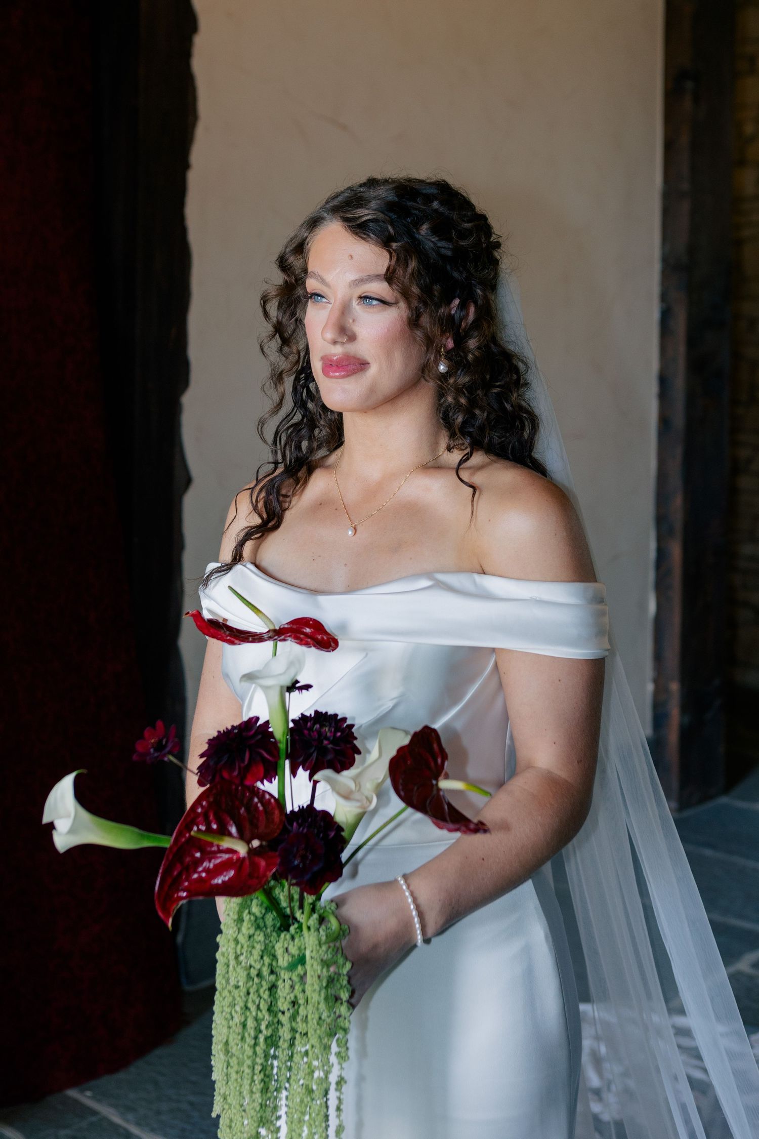 A bride in a white off-shoulder dress holds an elegant bouquet of dark red and white flowers with cascading greenery.
