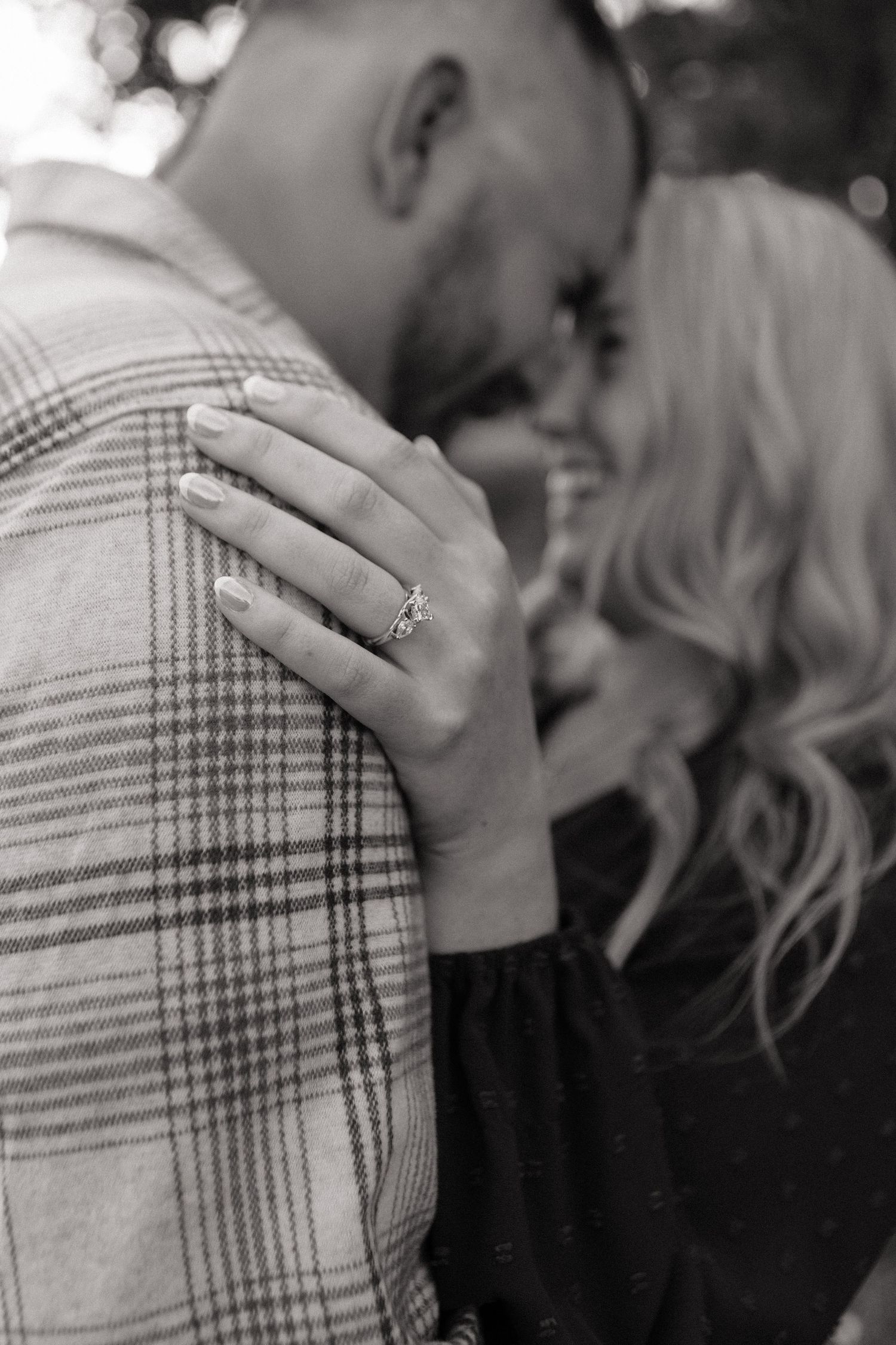Black and white photo of a couple embracing, woman's hand showing engagement ring on man's shoulder.