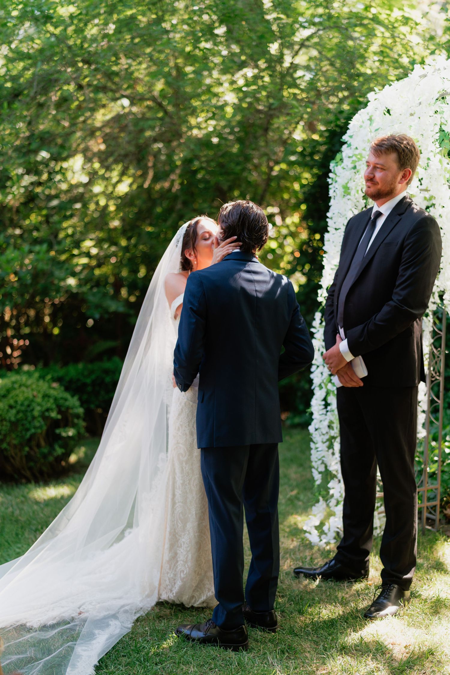 Bride and groom share first kiss at outdoor wedding altar adorned with white flowers while officiant watches.