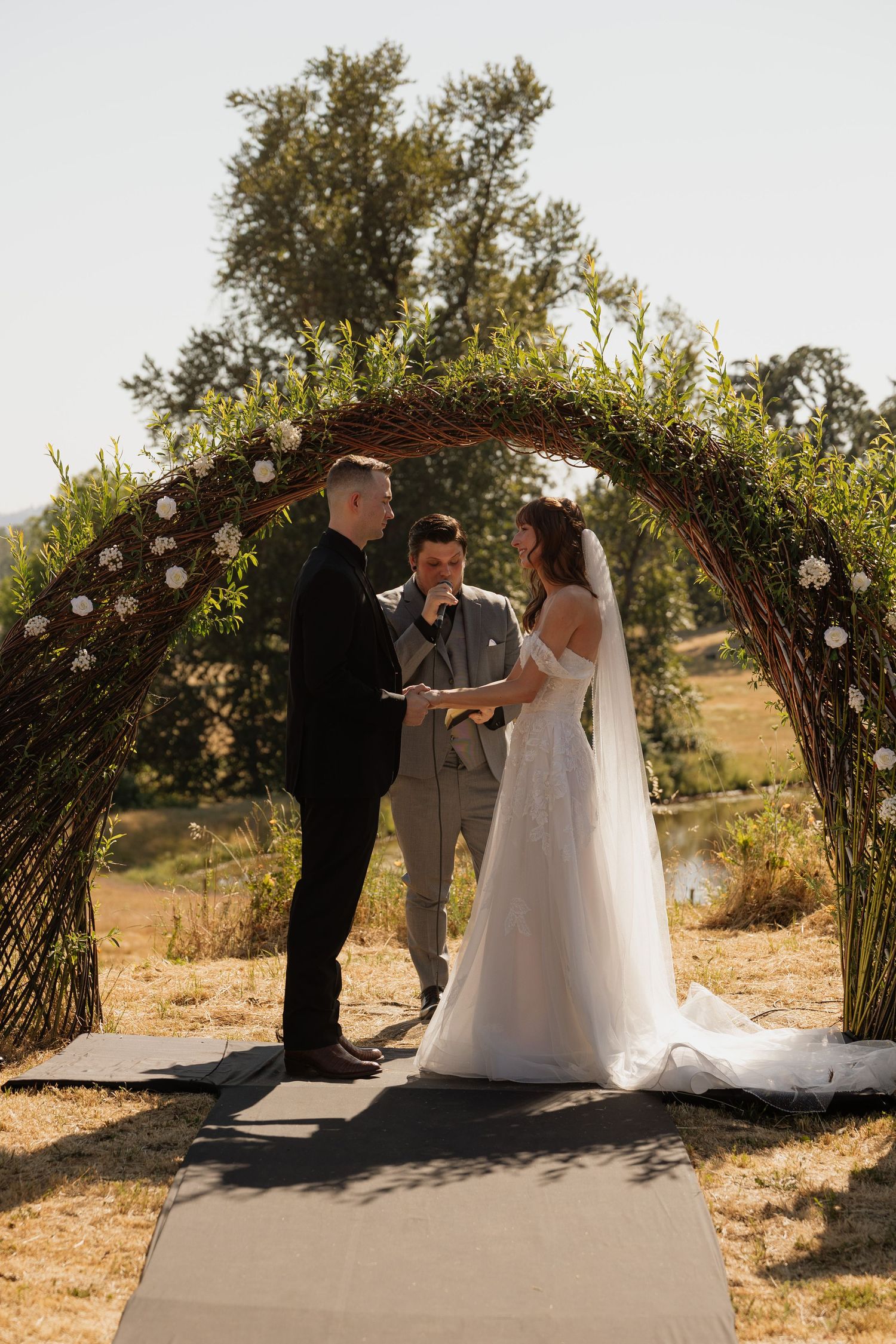 Bride and groom exchange vows under a rustic floral arch outdoors with a officiant during a sunny wedding ceremony.
