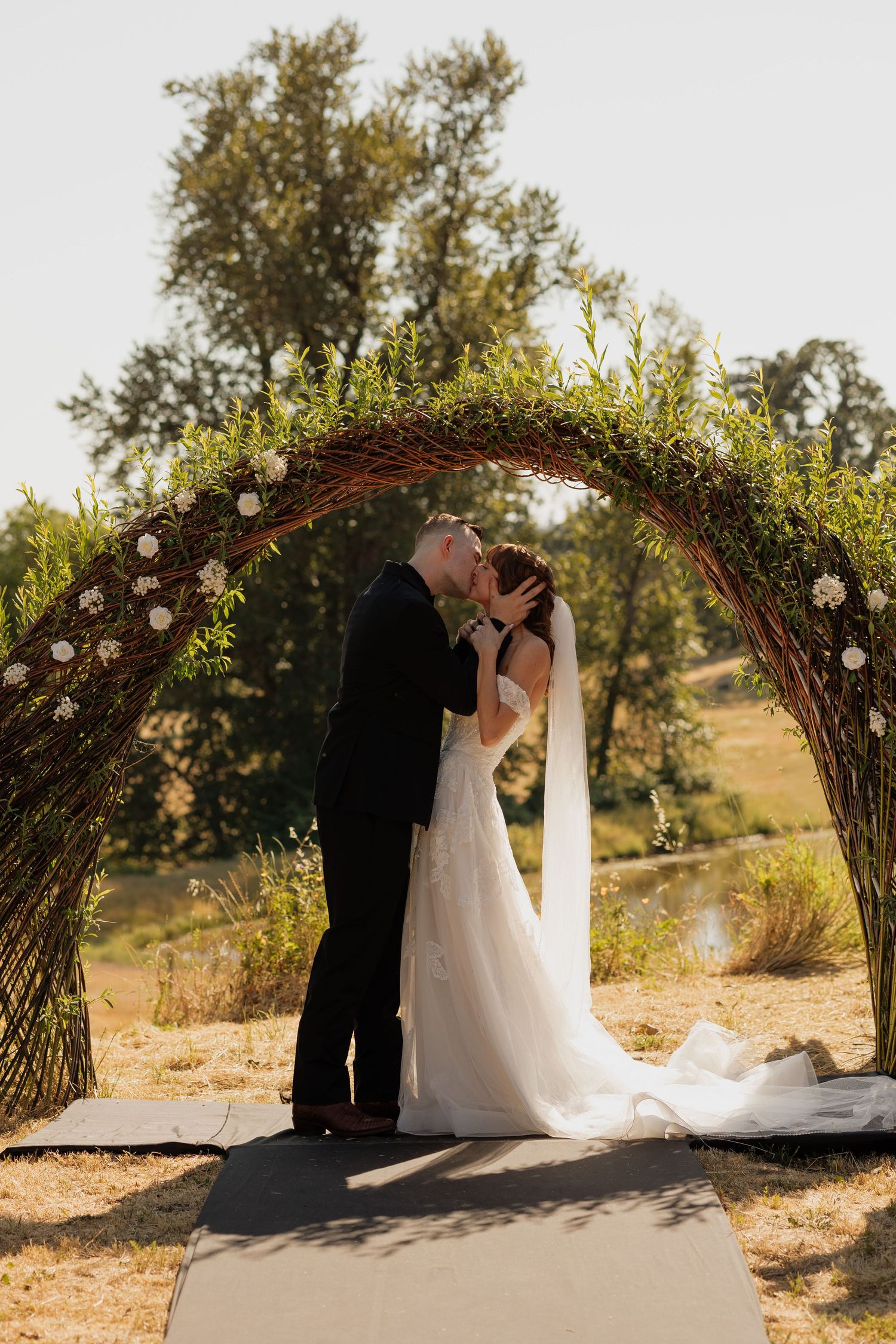 Bride and groom share first kiss under a rustic floral arch at an outdoor wedding ceremony.