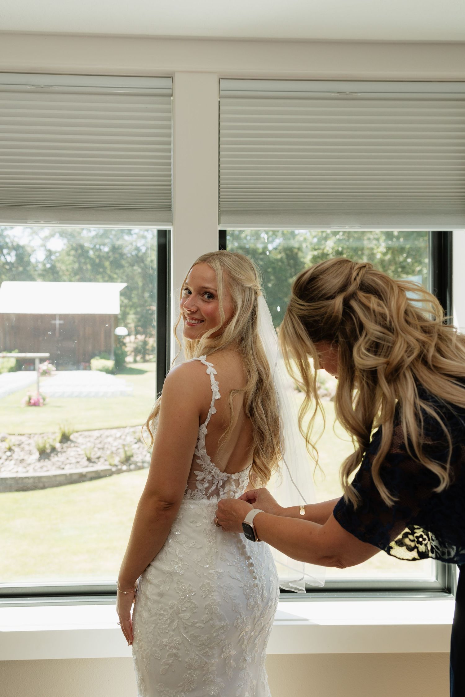 Bride in lace wedding gown smiling as bridesmaid buttons her dress near a bright window.