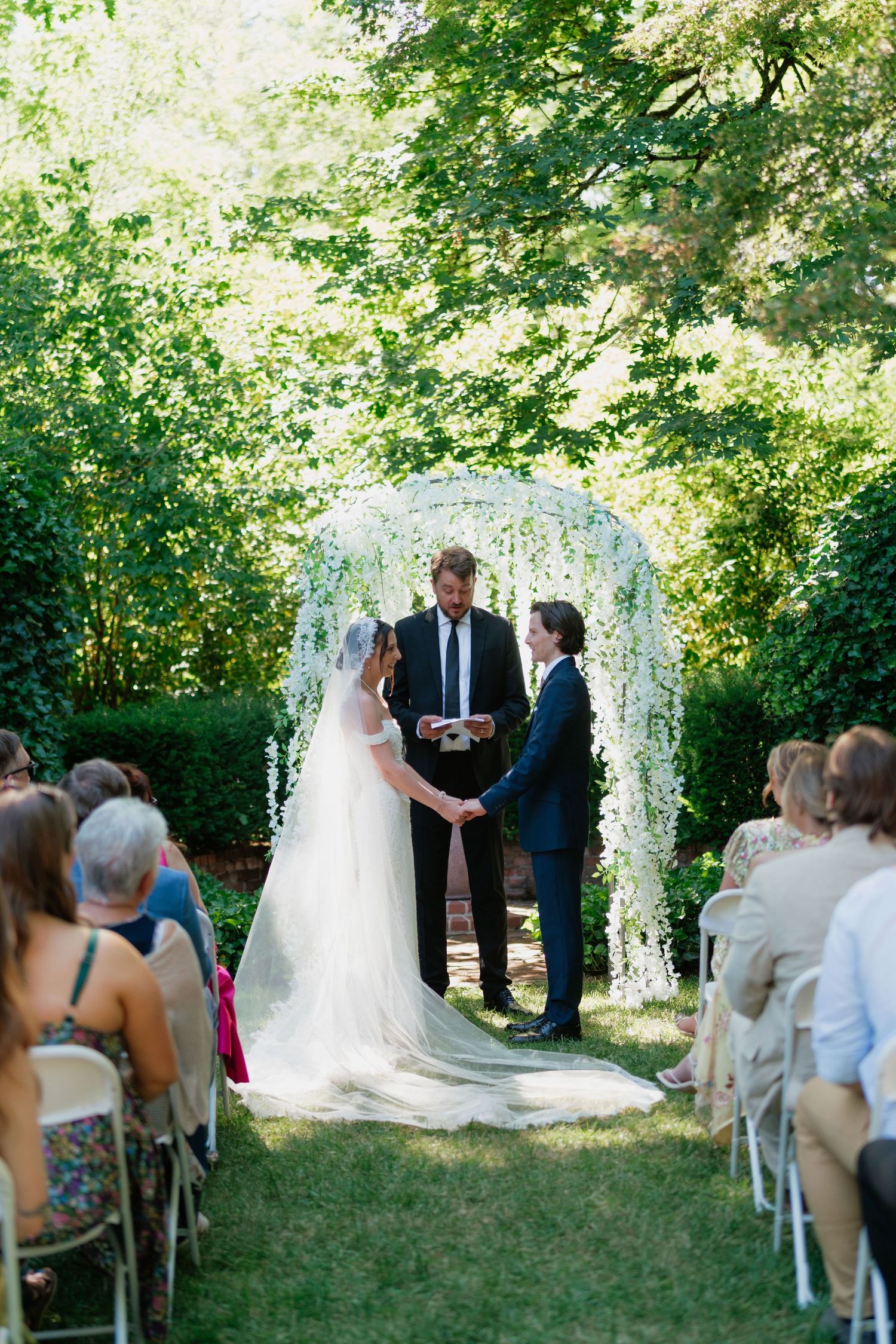 Bride and groom exchanging vows at outdoor garden wedding ceremony under white floral arch with seated guests.