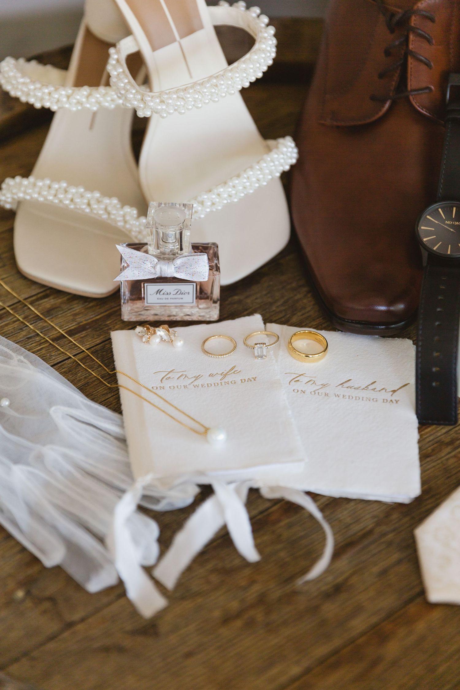 Wedding details including pearly white bridal sandals, wedding rings, perfume bottle and invitation on wooden surface.