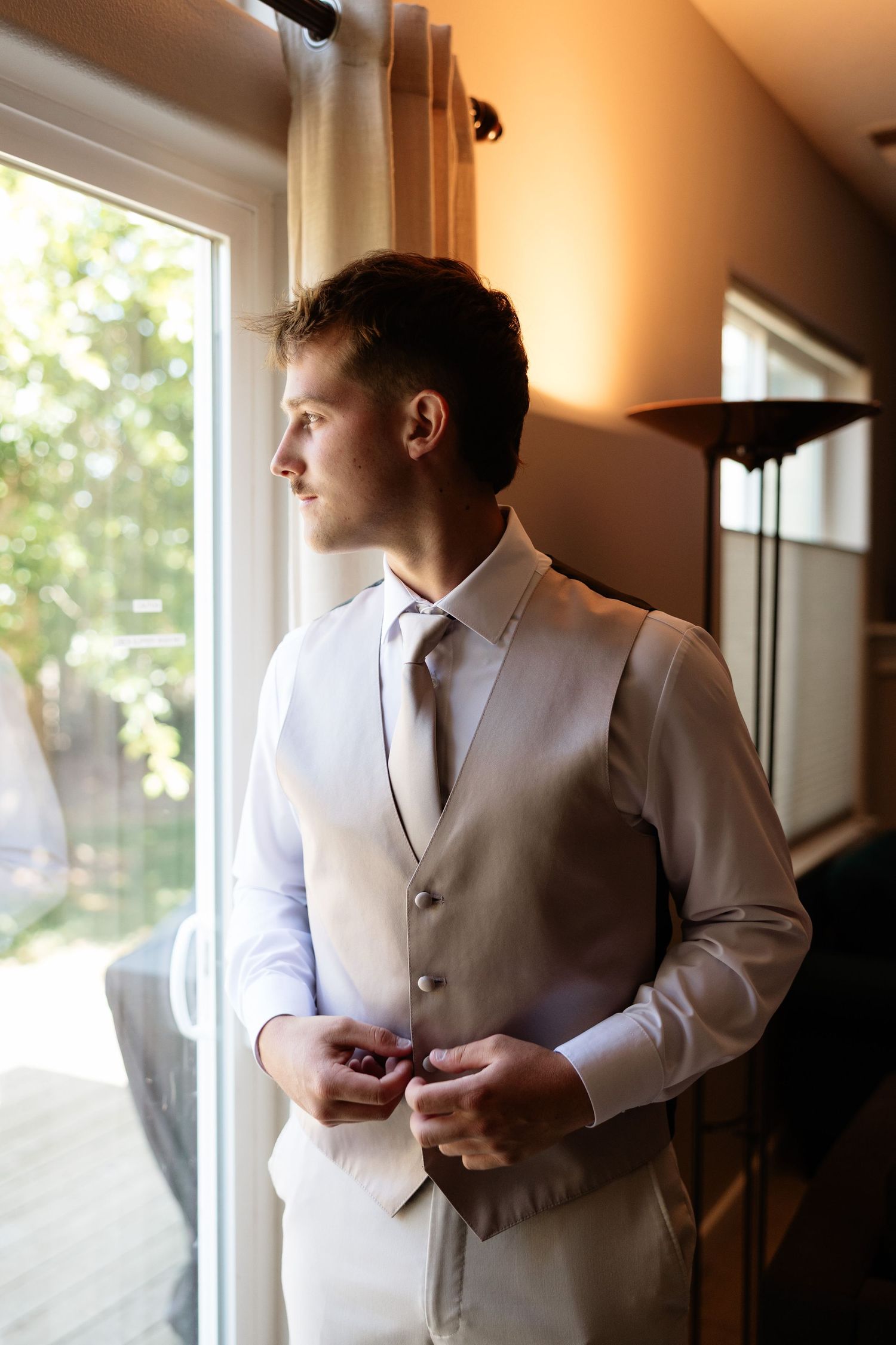 Handsome groom in beige vest and tie gazing out window while buttoning cufflinks before wedding.