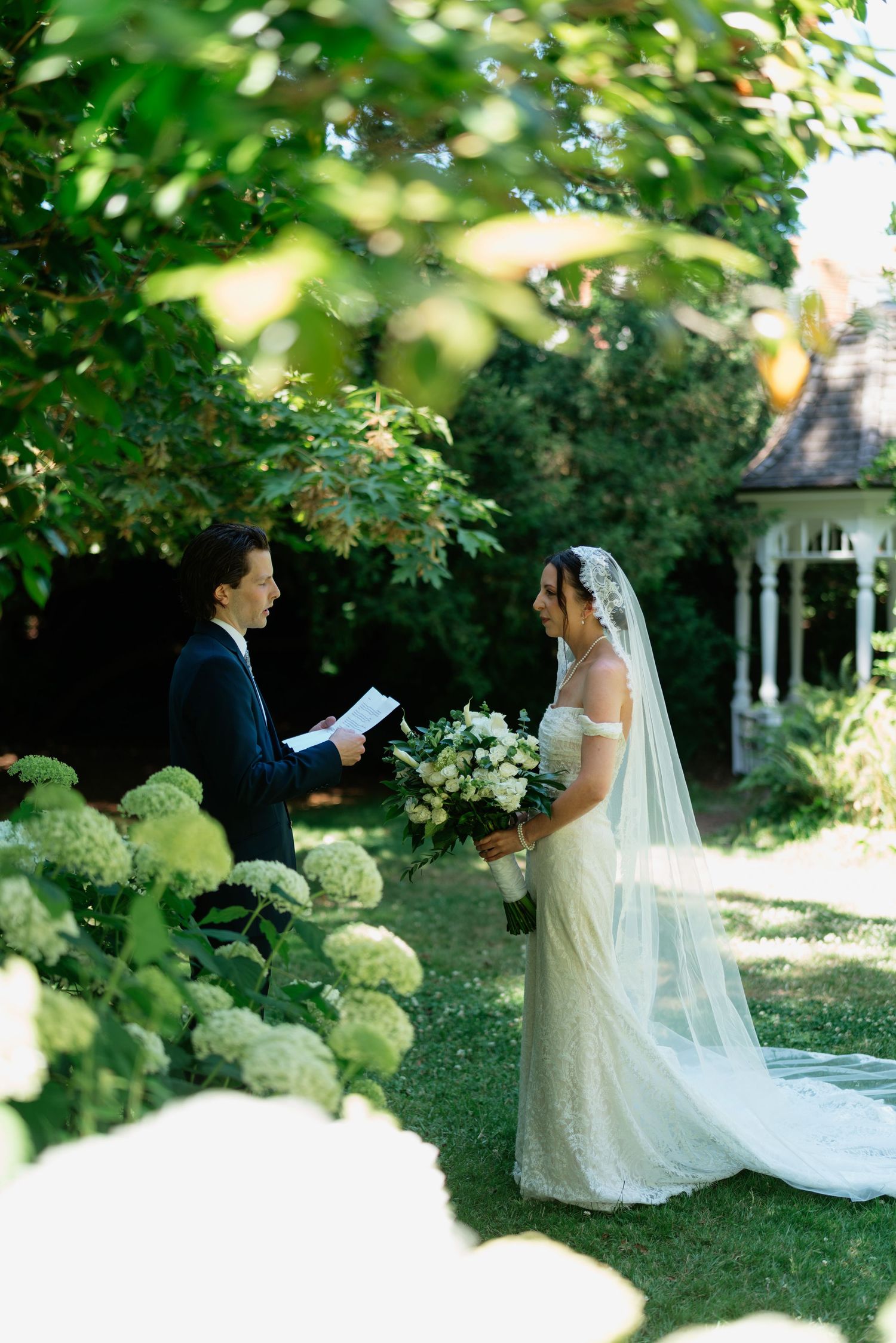 Bride in lace gown with veil holds bouquet as groom reads vows in lush garden ceremony near white gazebo.