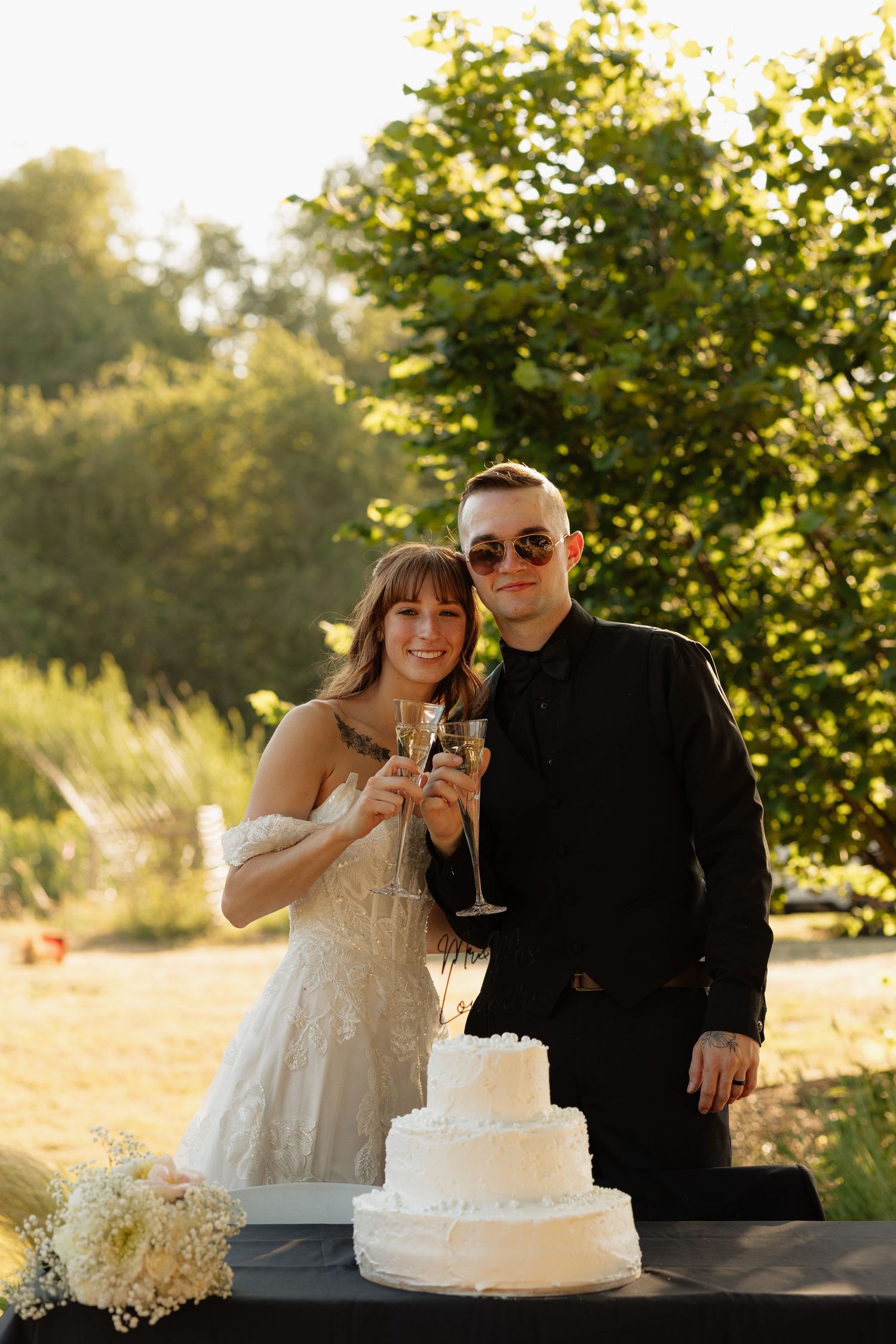 Bride and groom toasting with champagne glasses beside a three-tier white wedding cake outdoors in golden sunlight.