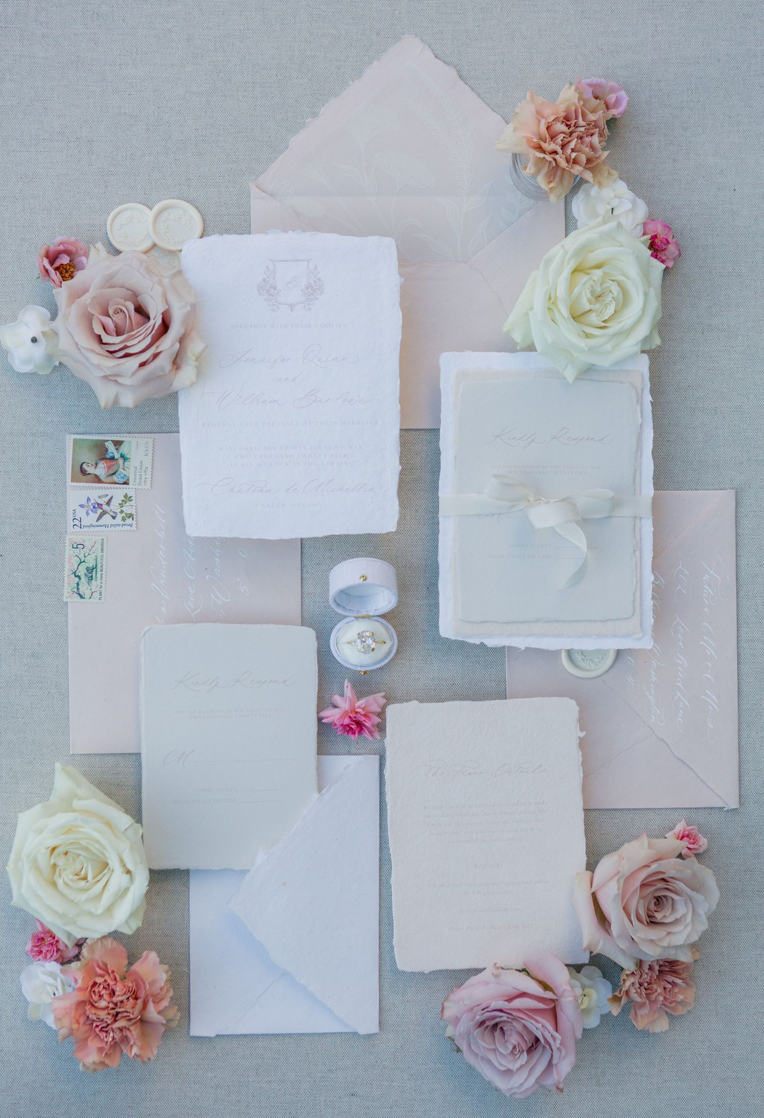 Flatlay of white wedding stationery with delicate pink and cream roses, vintage stamps, and a ring box on gray background.