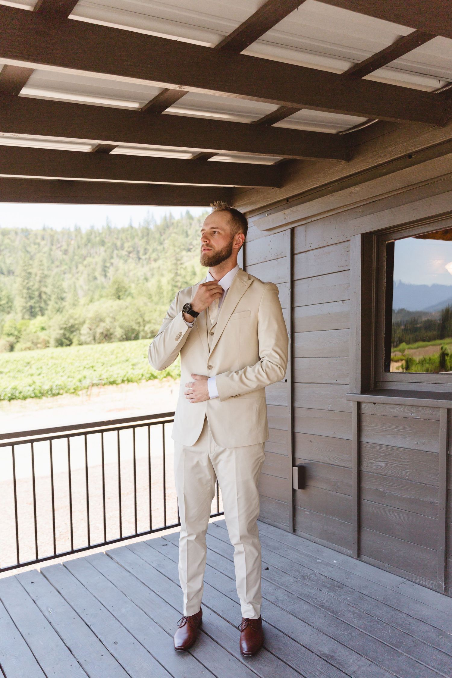 A person in a cream colored suit adjusts their tie while standing on a deck overlooking scenic mountain views.