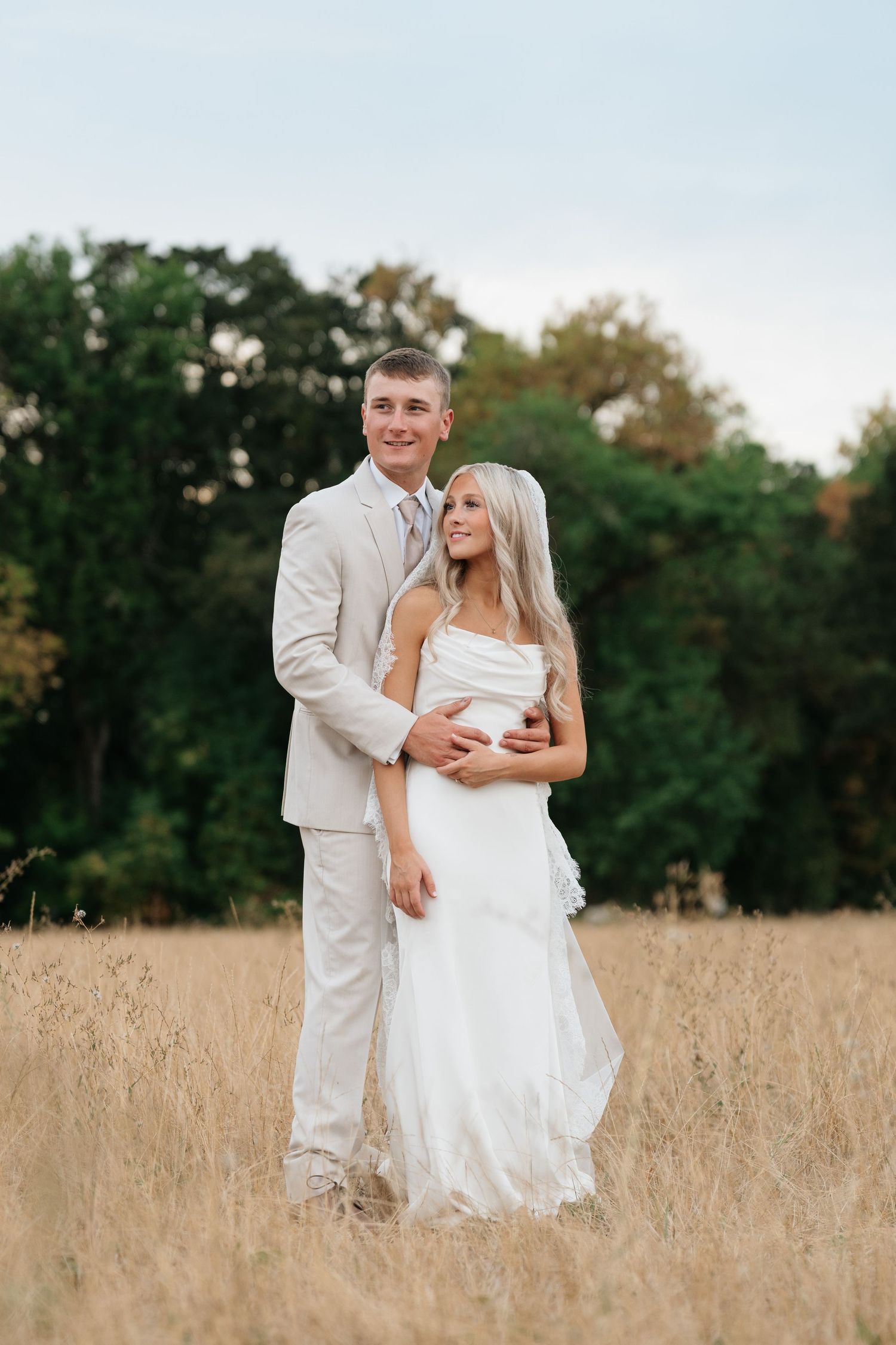 Bride and groom embracing in golden field wearing white wedding dress and beige suit with trees in background.